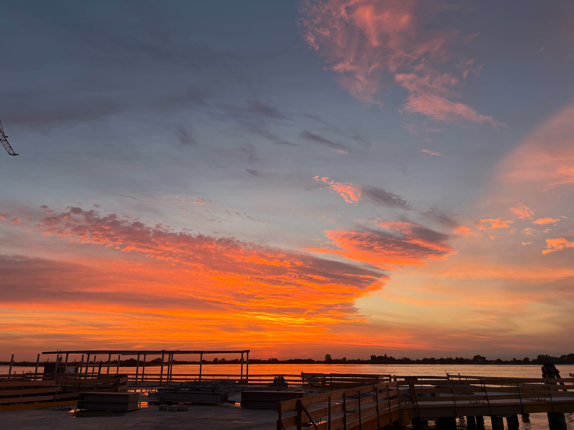 The image shows a sunset over the water in Punta Sabbioni, Italy. The sky is filled with vibrant orange and pink clouds. In the foreground, there is a construction site with wooden barriers and building materials. A crane is visible in the upper left corner of the image. The water is calm, and a distant shoreline with trees and buildings can be seen. A lone figure is standing on the pier in the lower right corner.