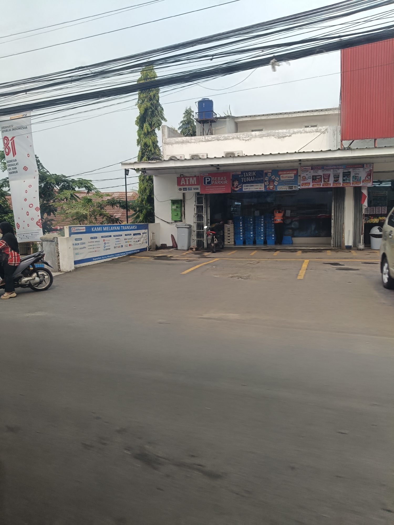 The image captures a street view in Saruni, Indonesia, focusing on an Alfamart convenience store and its surrounding area. In the foreground, a gray asphalt road with yellow parking lines is visible, suggesting a parking lot. To the left, a person wearing a black hijab and a red and black striped shirt is seated on a motorcycle.
The Alfamart store is the central subject, with its entrance open, revealing shelves stocked with various products. A banner above the entrance reads "KAMI MELAYANI TRANSAKSI" (We Serve Transactions) and lists various payment methods. Above this, prominent signage indicates "ATM" and "BEBAS PARKIR" (Free Parking). To the right of the store entrance, a beige car is partially visible.
The building features a white facade with a red section on the far right. A blue water tank is placed on the roof. A tall, slender green tree stands to the left of the building, adding a natural element to the urban landscape. Overhead, a dense network of black power lines crisscrosses the sky. A white banner with red text and circular designs is displayed on a pole to the left of the store, featuring "DIRGAHAYU REPUBLIK INDONESIA" and the number "81". The overall atmosphere is that of a typical commercial area in an Indonesian city.