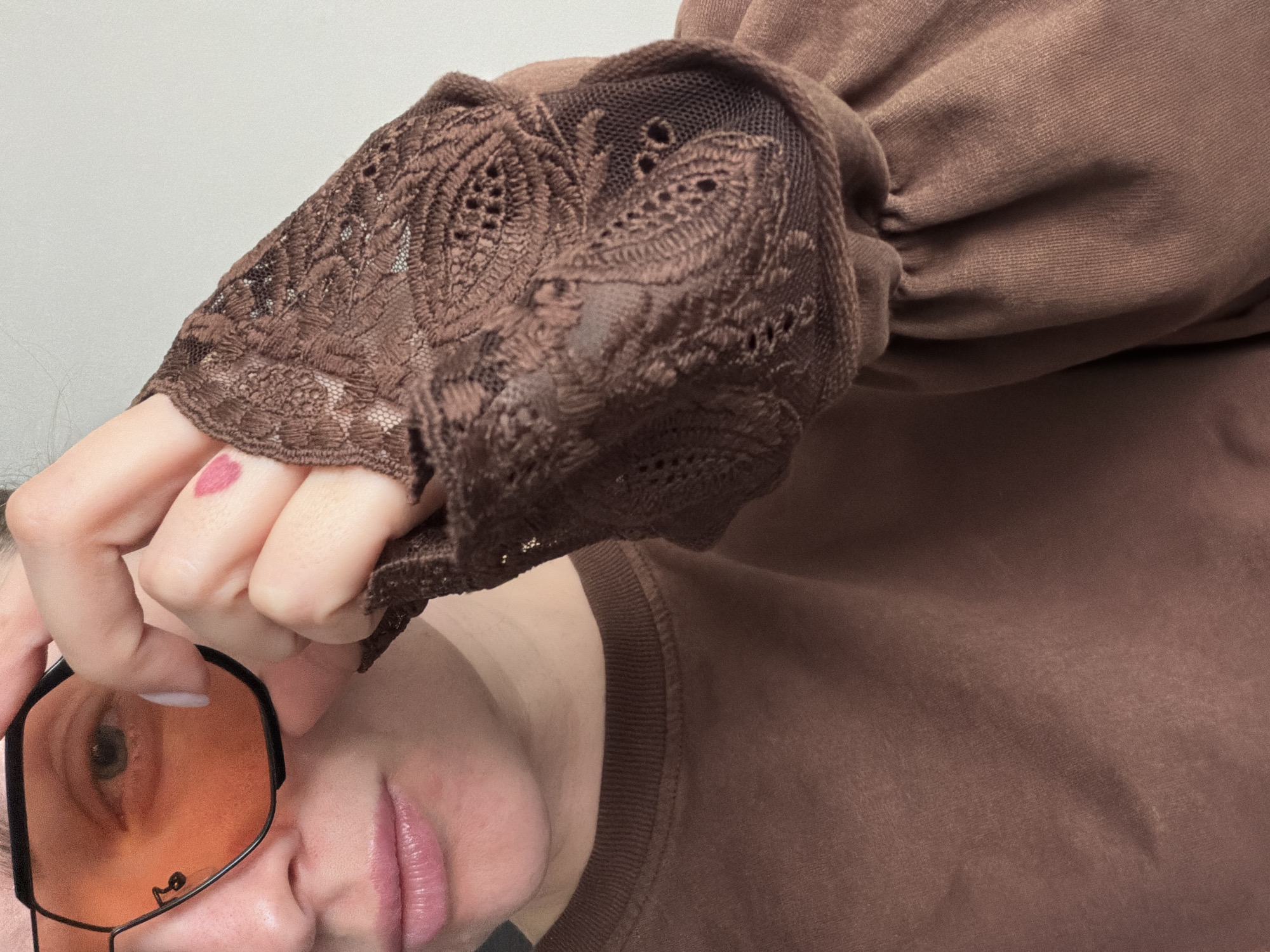 The image shows a person holding glasses with orange lenses in front of their face. They are wearing a brown long-sleeved top with lace detailing on the sleeves. The background is a plain, light-colored wall. There are no indications of the city of Moscow, Russia, or any other specific location.