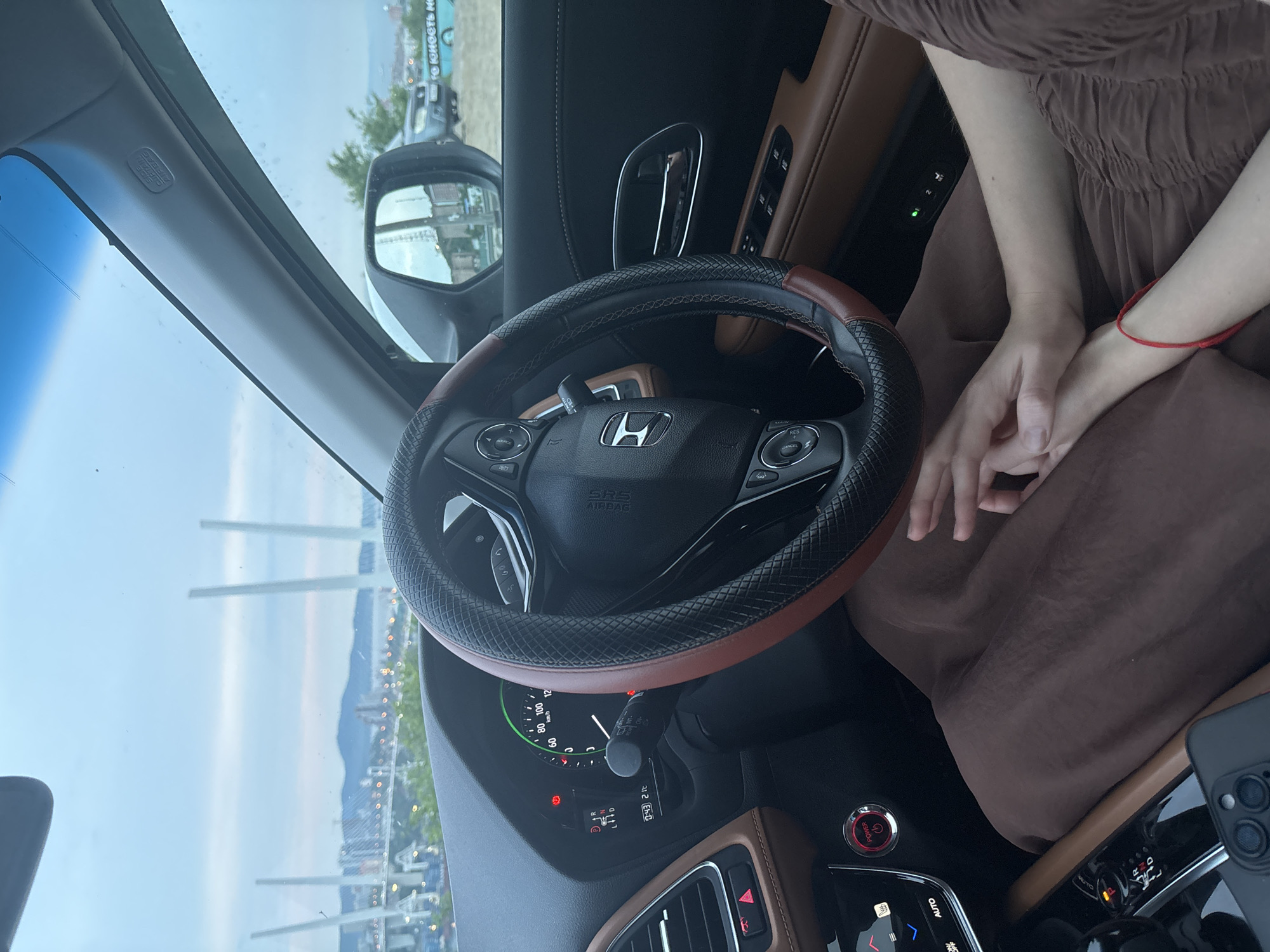 The image shows the interior of a car with a view of the Golden Horn Bay and the Golden Bridge in Vladivostok, Russia, through the windshield. The dashboard, steering wheel, and a portion of the passenger's lap are visible. Two people's hands are intertwined in the foreground, with one person wearing a red bracelet. The car appears to be parked, with the engine off. The sky is overcast.