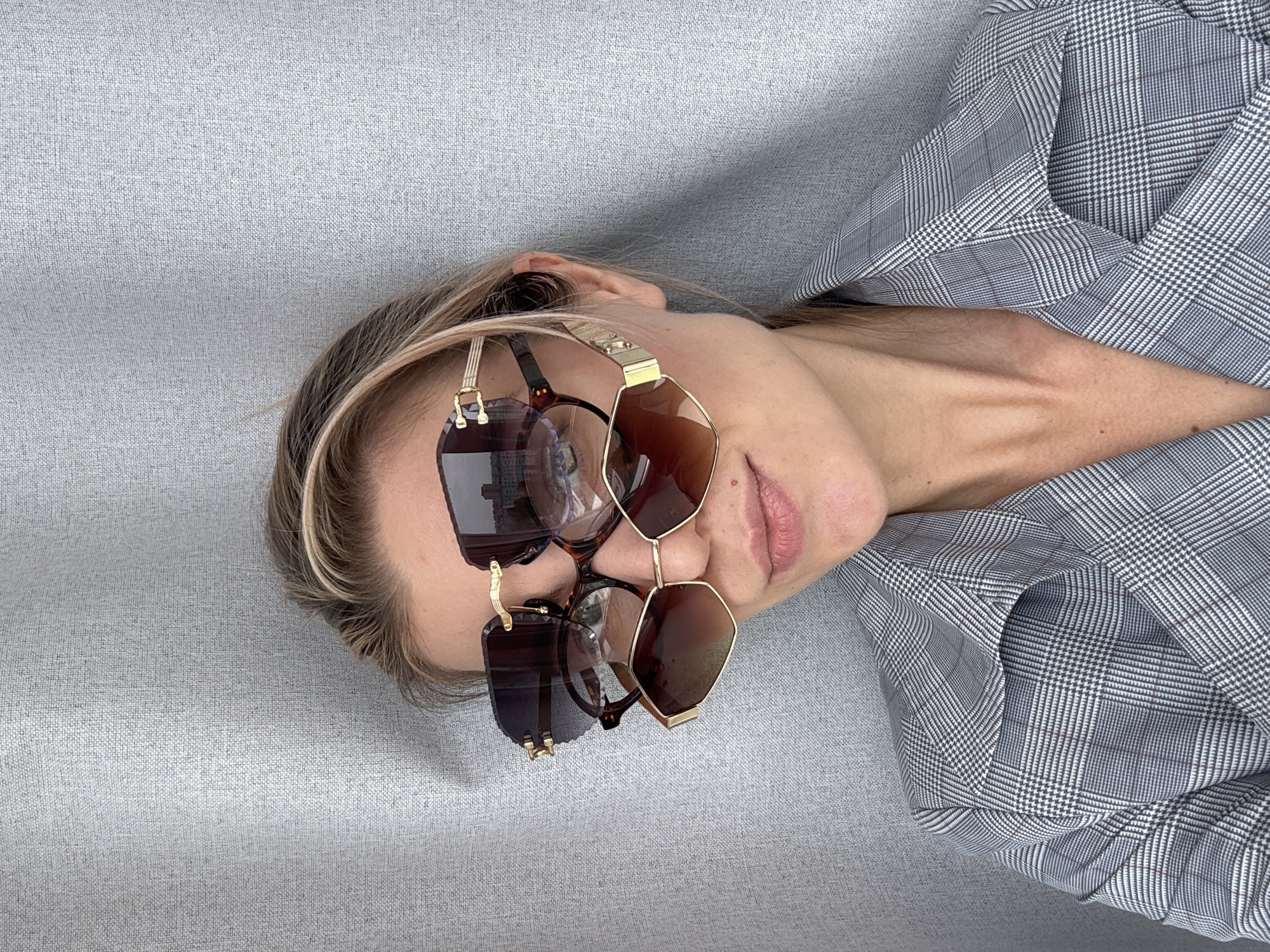 A woman lies on a grey textured surface, possibly a couch, wearing a grey and white checkered blazer. She is facing the camera, her head tilted to the right. Two pairs of sunglasses are balanced on her head and nose. The sunglasses have a gold frame and brown lenses. The woman has fair skin, blonde hair, and pink lips. The location is Unknown, Unknown.