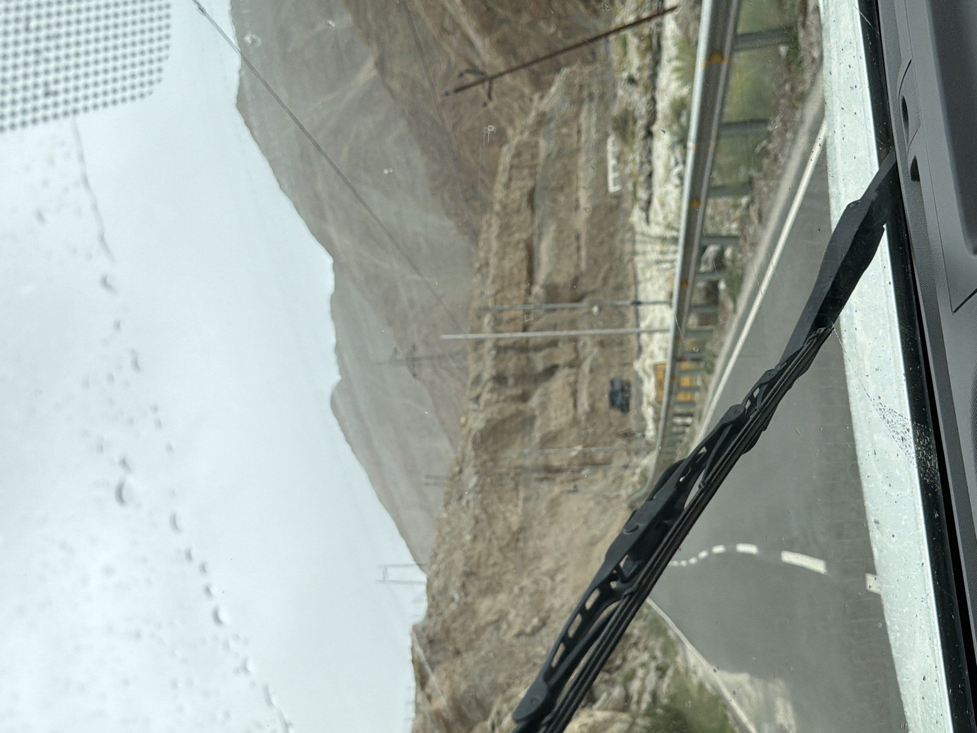 This image, taken from inside a vehicle, shows a view through a wet windshield of a road passing through a mountainous, rocky terrain. The road is paved and has white lane markings. Guardrails are visible along the side of the road. In the background, steep, eroded hillsides rise up, with evidence of some construction or power lines. A small dark vehicle is visible further down the road. The sky is overcast and appears to be raining, as indicated by the water droplets on the windshield. The location is identified as Ikpadok, India.