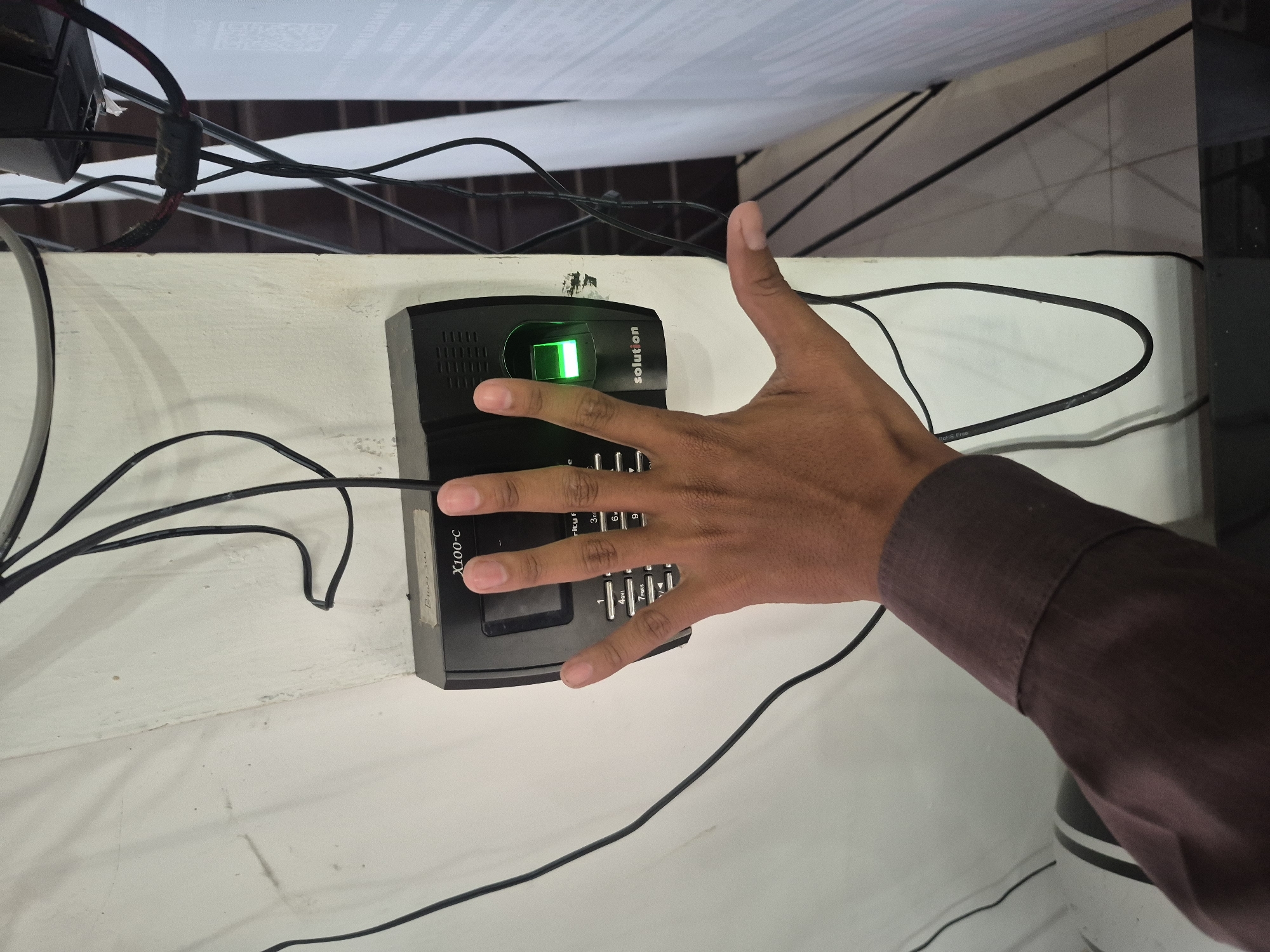 A person is using a fingerprint scanner. The scanner, labeled "X100-C Solution," is mounted on a white wall, surrounded by black electrical wires. A green light indicates the scanner is active. The person's right hand is positioned over the scanner, with the index finger placed on the fingerprint reader. The person is wearing a dark-colored long-sleeved shirt. The location is identified as the city of Lhokseumawe, Indonesia.