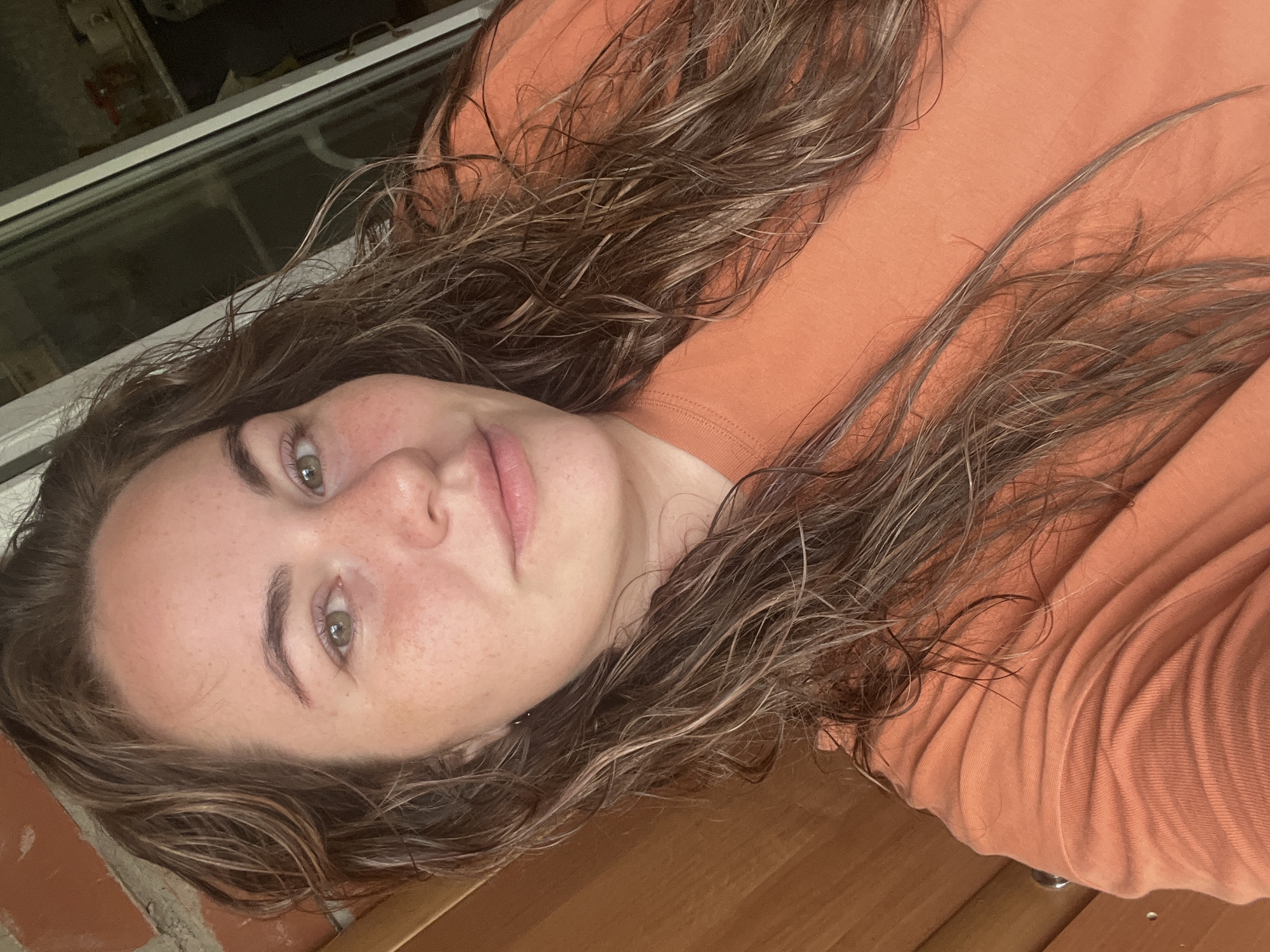 A close-up selfie shows a young woman with long, wavy brown hair and fair skin with freckles. She is wearing an orange t-shirt. Her eyes are green, and she has a slight smile. The background appears to be indoors with wooden paneling and a window. The image is taken in Chaikovsky, Russia.