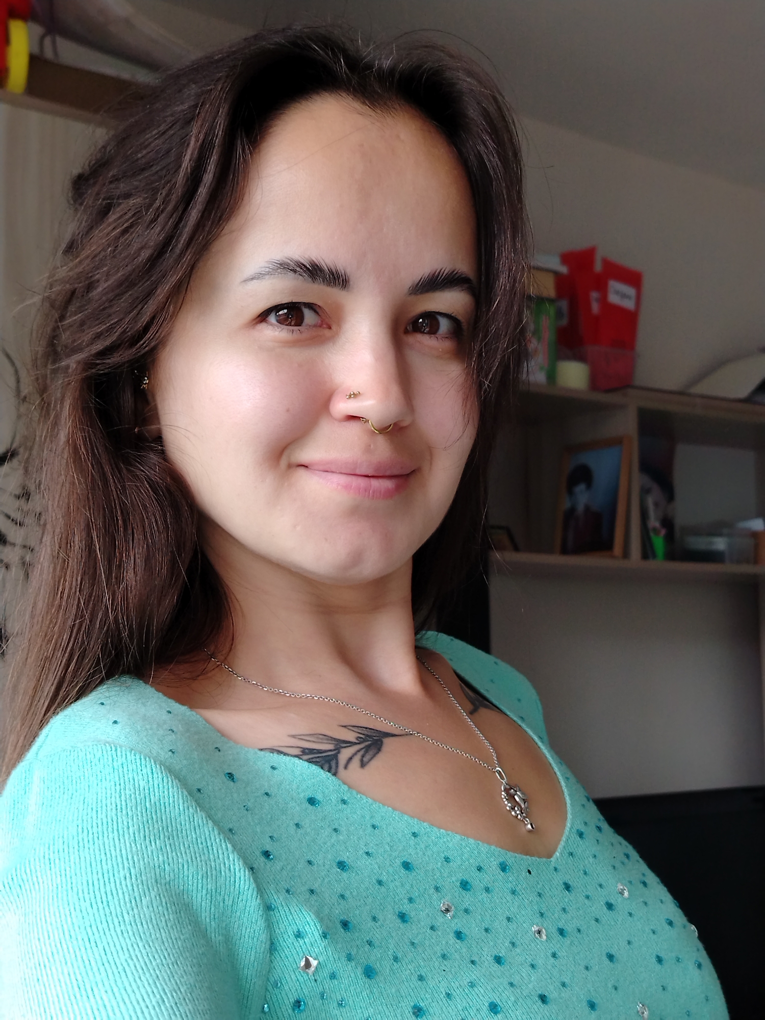 The image shows a close-up of a young woman with long, dark hair and brown eyes. She is wearing a light teal sweater adorned with small rhinestones. She has a septum piercing and a necklace with a pendant. A tattoo is visible on her chest. The background appears to be a room with shelves holding various items, including a framed picture. The lighting suggests an indoor setting, likely a home. There is no indication of the city of Noviy, Russia.
