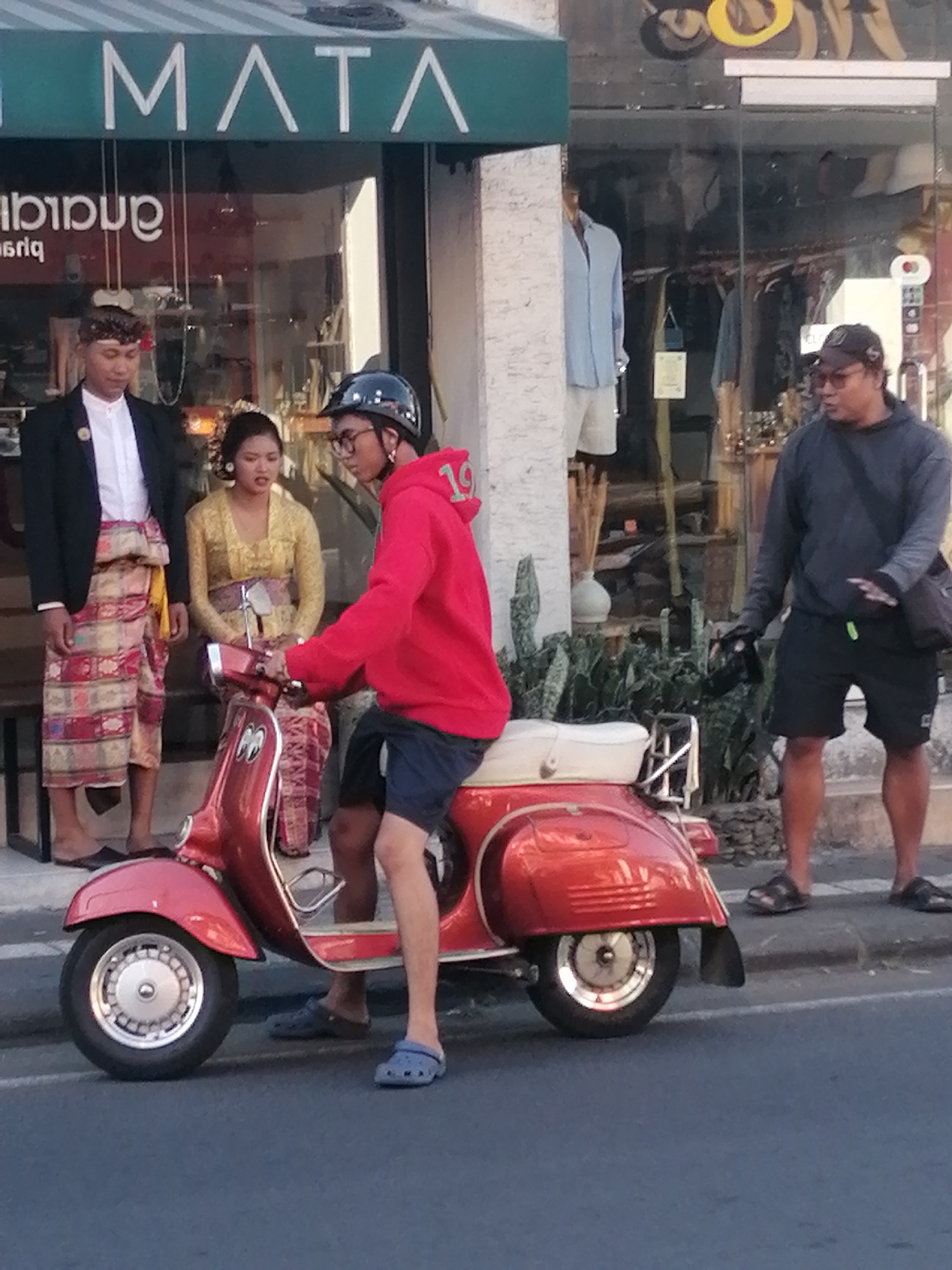 A man in a red hooded sweatshirt with a black helmet and glasses rides a red scooter. He is wearing blue shorts and blue croc-style sandals. To his left, a woman in a yellow traditional outfit stands next to a man in a black jacket and a patterned sarong. They are standing in front of a store with the word "MATA" visible on the awning. Behind them, a man in a dark grey hooded sweatshirt and black shorts stands with a black bag slung over his shoulder. The scooter is parked on a street in Ubud, Indonesia, with a sidewalk and plants visible in the background.