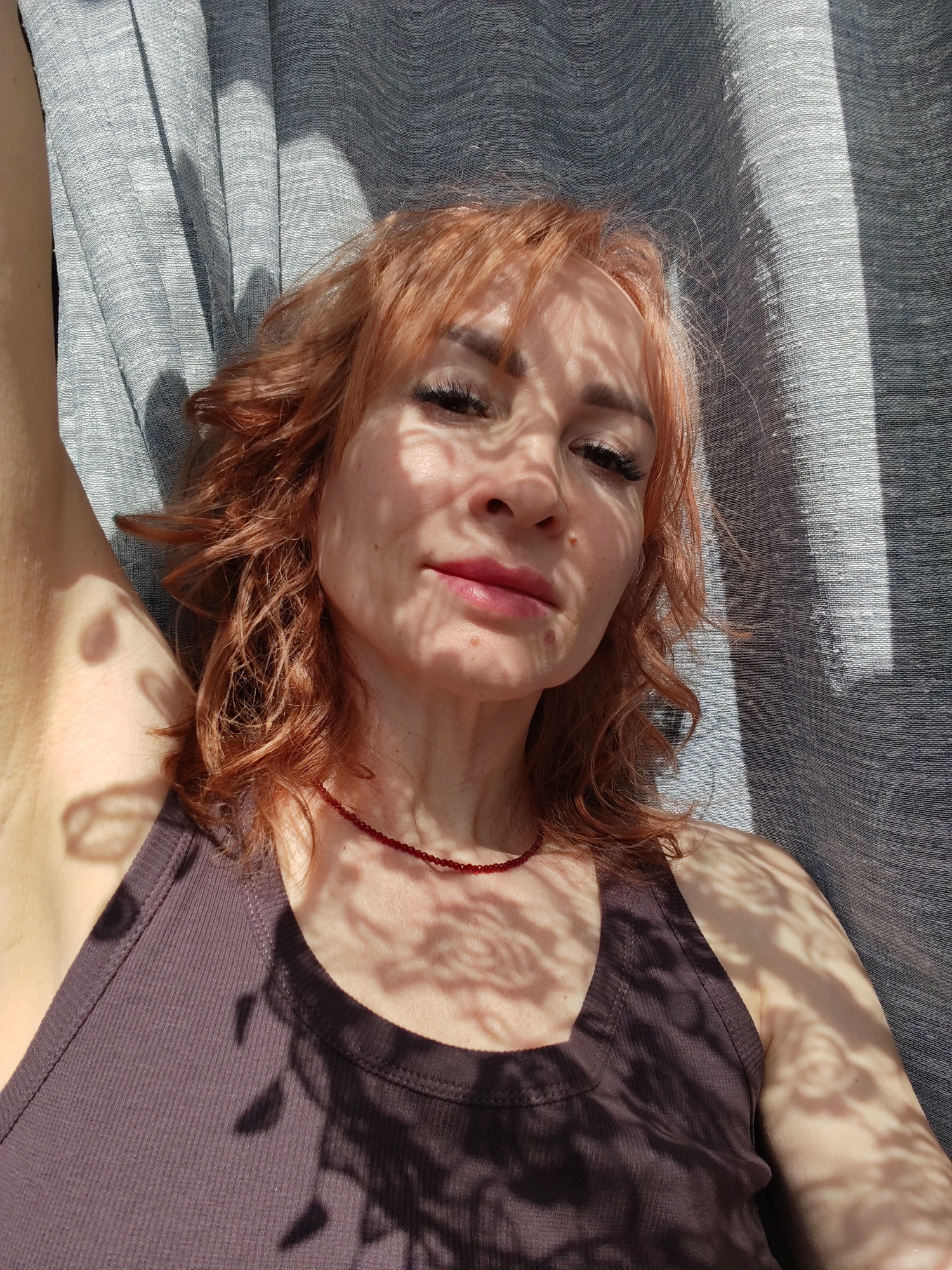 A woman with reddish-blonde hair, wearing a dark brown tank top and a red bead necklace, is lying down in an indoor setting. Her left arm is raised. Light and shadow patterns are cast across her face, body, and the gray fabric behind her. The location is Старое Михайловское, Russia.