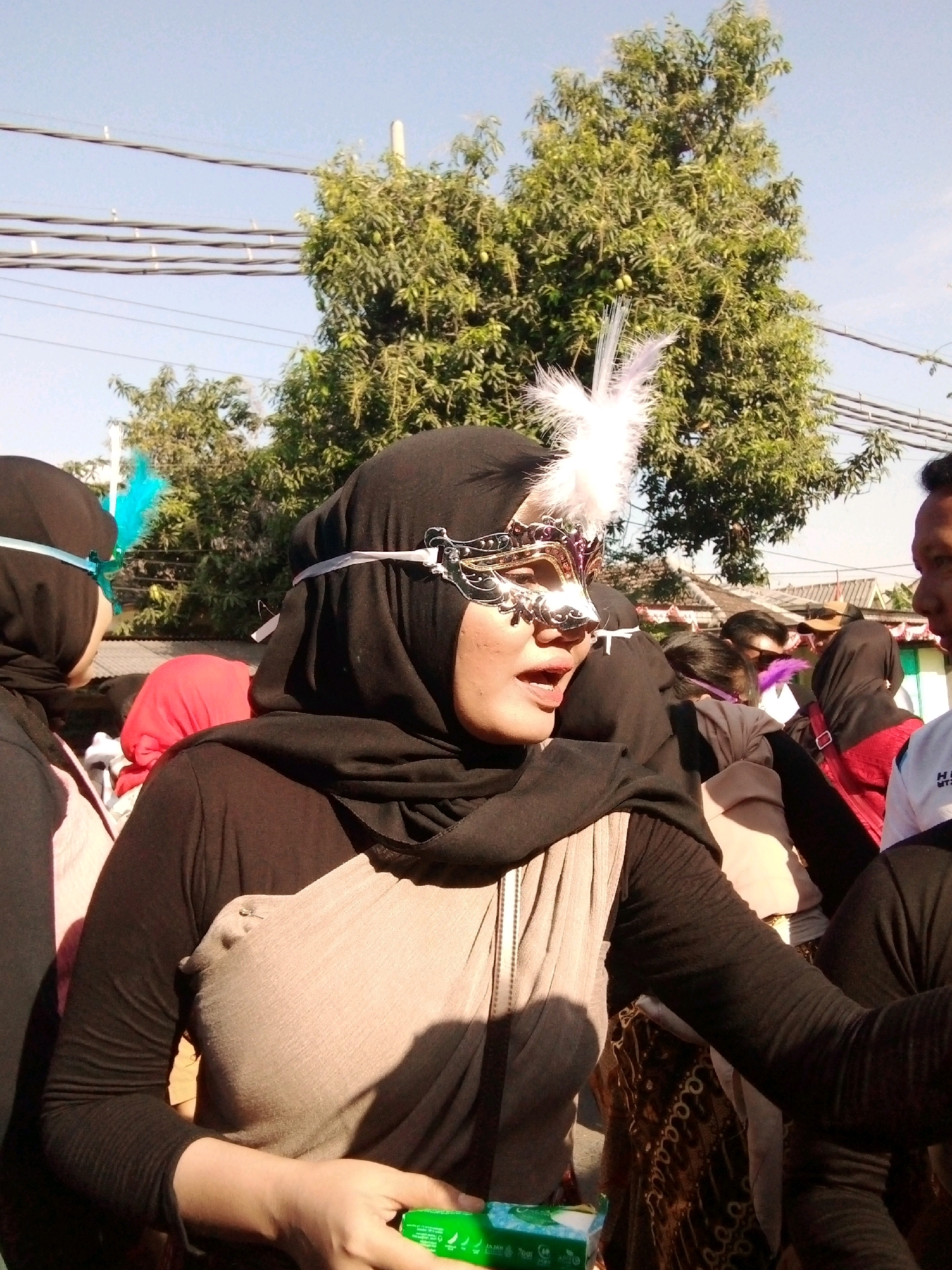 In Sumberwono, Indonesia, a woman wearing a black hijab and a masquerade mask with white feathers on it is visible in the foreground, speaking. She is wearing a light brown scarf draped over her shoulders and a black long-sleeved shirt. Behind her, other people are present, some wearing hijabs. Trees and overhead wires are seen in the background under a clear sky. The scene appears to be outdoors, possibly during a daytime event or parade.