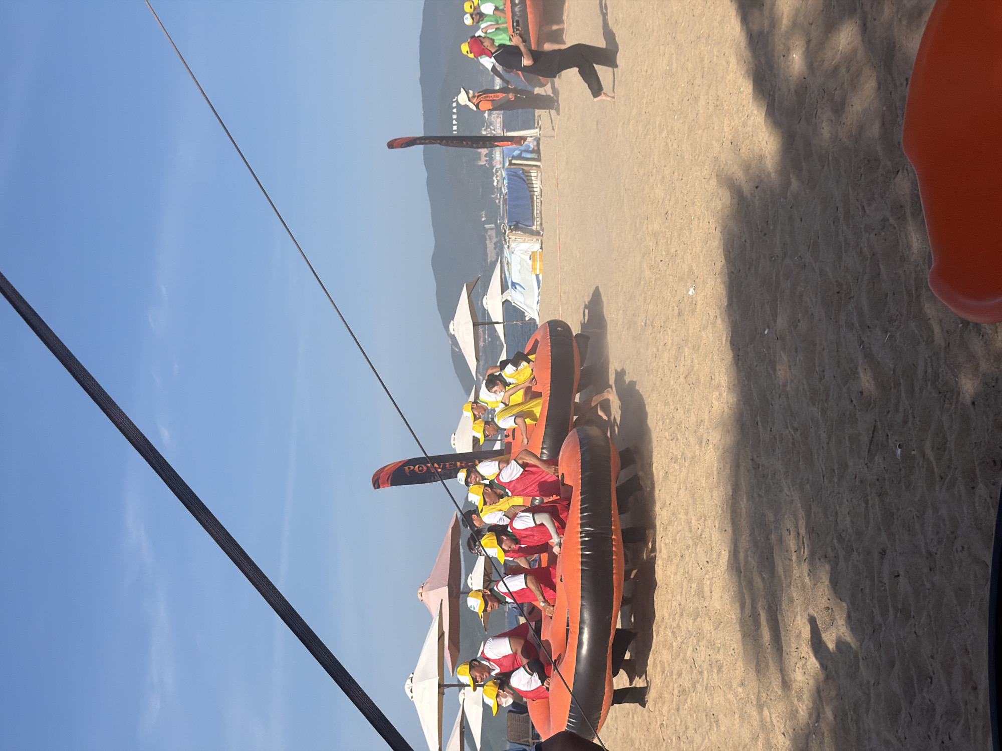 The image depicts a group of people participating in a beach activity in Nha Trang Ward, Vietnam. The foreground is dominated by a sandy beach with some shadows cast from trees. In the middle ground, several individuals are seen running and pushing large, inflatable orange tubes on the sand. The people are wearing yellow caps and red vests, suggesting they are part of a team or organized event. Some of the inflatable tubes have black accents. In the background, there are several beach umbrellas, a body of water, and a hilly coastline with some buildings visible. The sky is clear and blue. There is a tall, black, diagonal rope or cable stretching across the upper portion of the image, likely part of a zipline or similar overhead structure. There are also flags planted in the sand, one of which has the word "POWER" visible. The overall scene conveys a sense of active recreation and community gathering on a sunny day.
