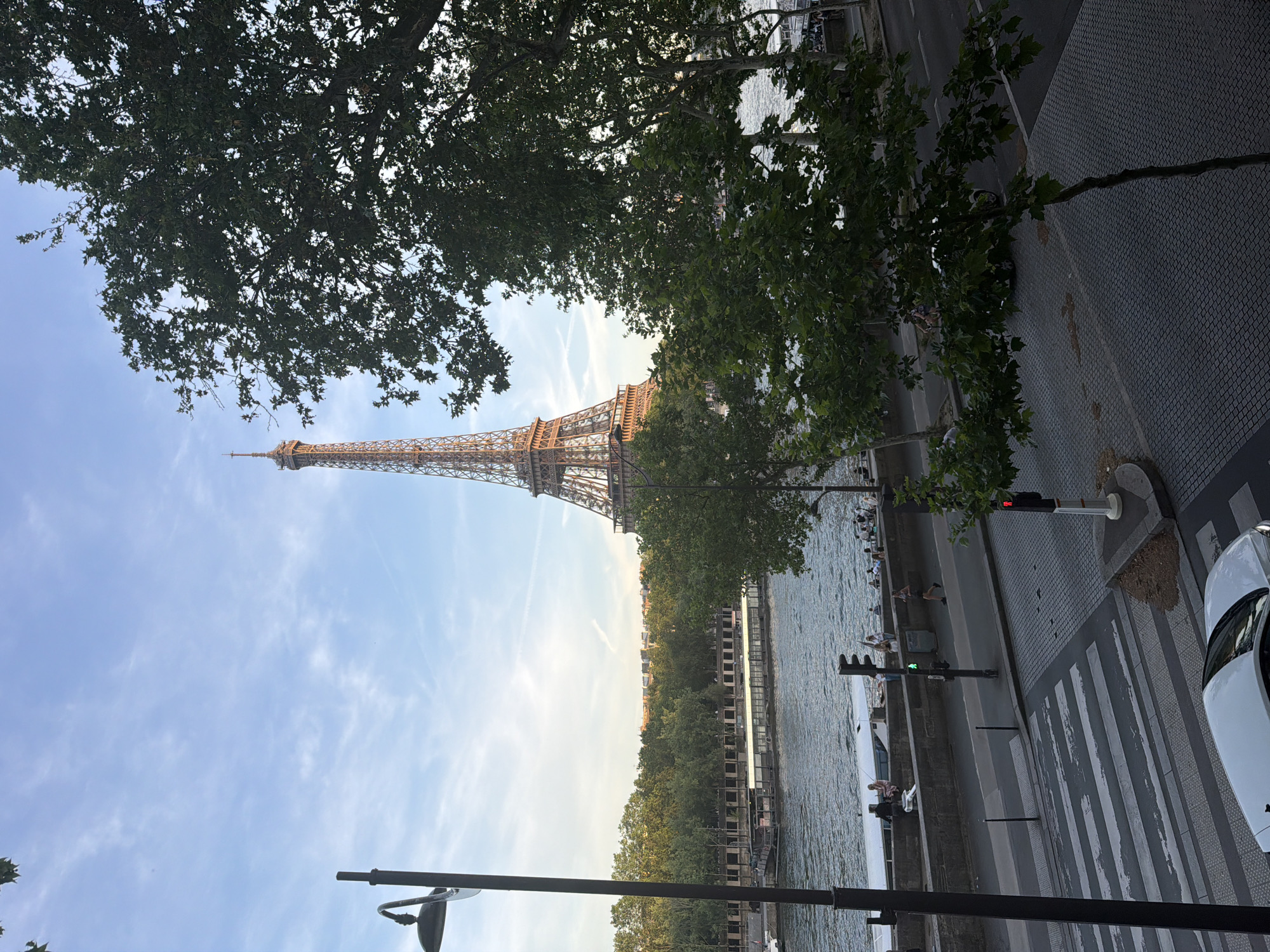 The image shows the Eiffel Tower in Paris, France, viewed from a street-level perspective with trees framing the tower. The scene includes the Seine River running alongside the road. People are seated near the riverbank. A pedestrian crosswalk and traffic light are visible on the side of the road, along with a car.
