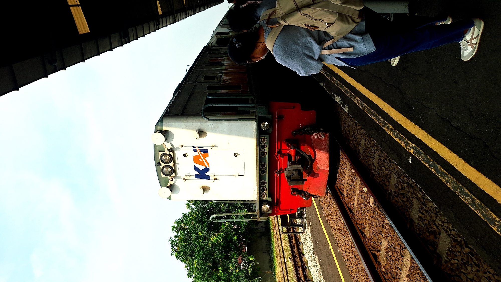 The image shows a train at a station in Karawang, Indonesia. A person wearing a gray sweatshirt, blue pants, and white sneakers stands on the platform. They have a backpack on. The train is predominantly white with a red front and the KAI (Kereta Api Indonesia) logo. The station platform has yellow lines marking the edge. Train tracks with gravel are visible beside the platform. Trees are visible beyond the tracks. The sky is clear and blue. The photo is taken at an angle.