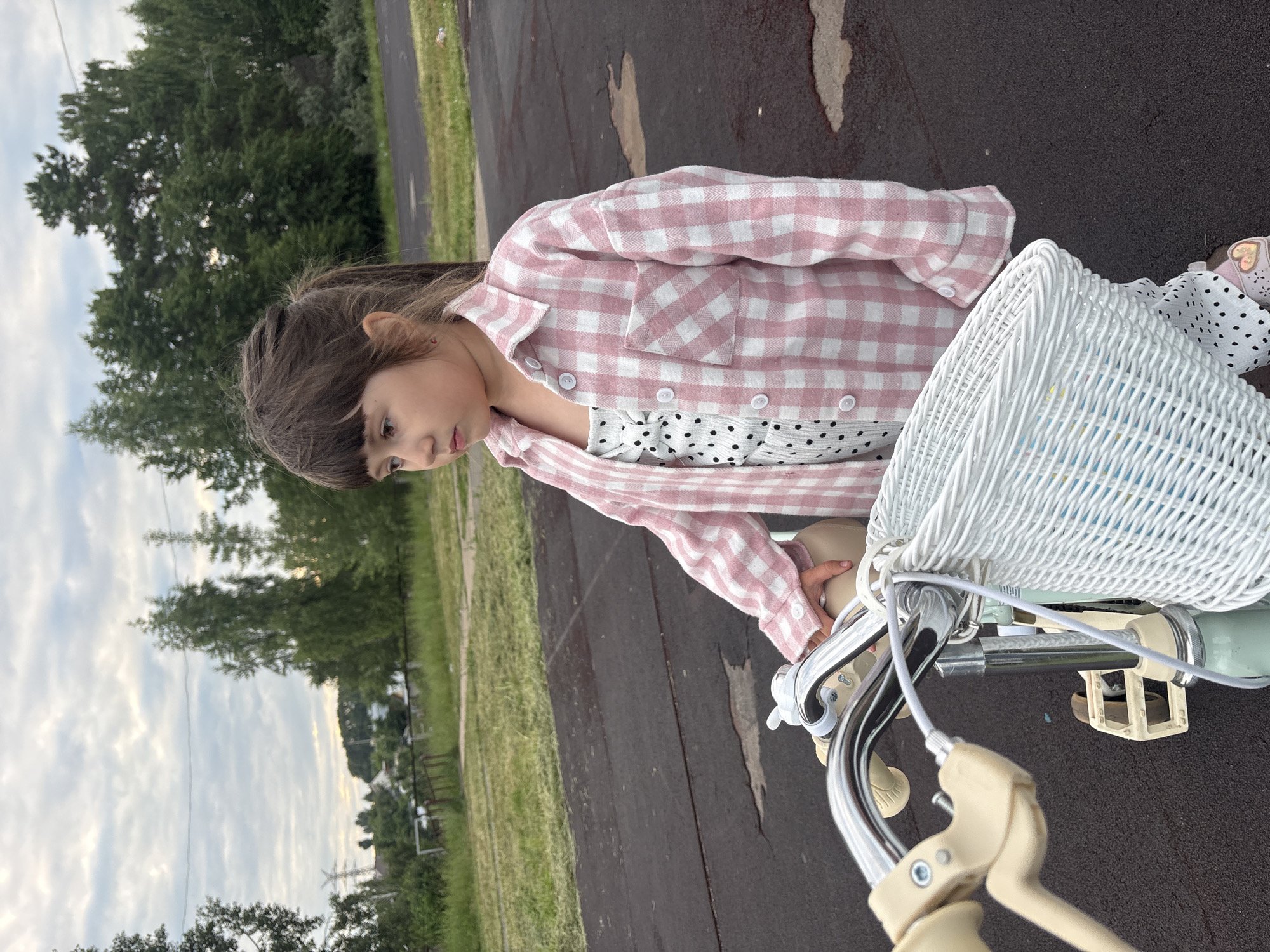 A young girl stands next to a light-colored bicycle on an asphalt surface, likely a park or playground area, in Chaikovsky, Russia. The girl is wearing a white and black polka dot jumpsuit with a pink and white plaid shirt over it. She has dark hair tied in a ponytail and is looking down at the bicycle. The bicycle is equipped with a white wicker basket and has light-colored handlebars and pedals. In the background, there are trees and a cloudy sky. The overall setting appears to be outdoors in a semi-rural or suburban environment.
