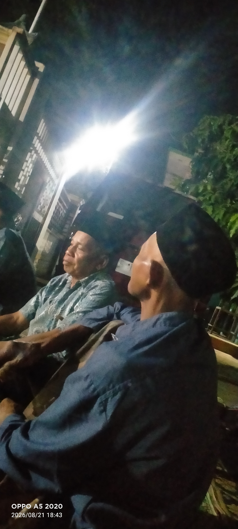 In Sowan Kidul, Indonesia, at night, two individuals are seated outdoors under a bright artificial light. The person on the left is an older man wearing a patterned blue shirt and a black peci (a type of cap). He is looking to his left. To his right, a second person is also seated, their face turned away from the camera, also wearing a black peci and a blue shirt. The environment appears to be near a building with a light-colored fence or wall. Trees are visible in the background. The bottom right corner of the image contains a timestamp and device information: "OPPO A5 2020 2026/08/21 18:43".