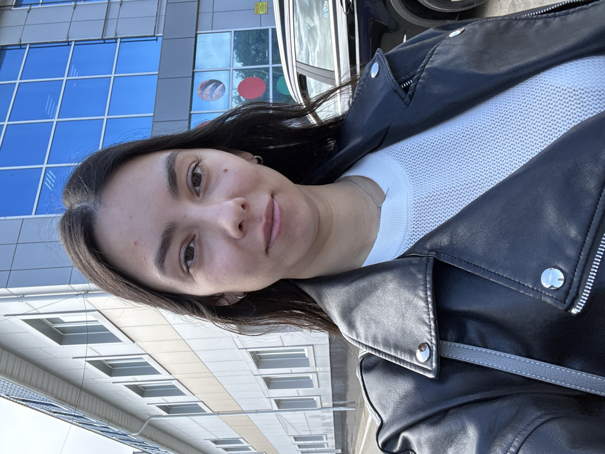 A person with dark hair and fair skin stands outside a building in what appears to be Chaikovsky, Russia. The person is wearing a white top and a black leather jacket. A building with blue windows and another with beige and white horizontal panels are in the background. A dark car is partially visible behind the person. Reflections of planetary or colorful shapes can be seen in one of the blue windows.