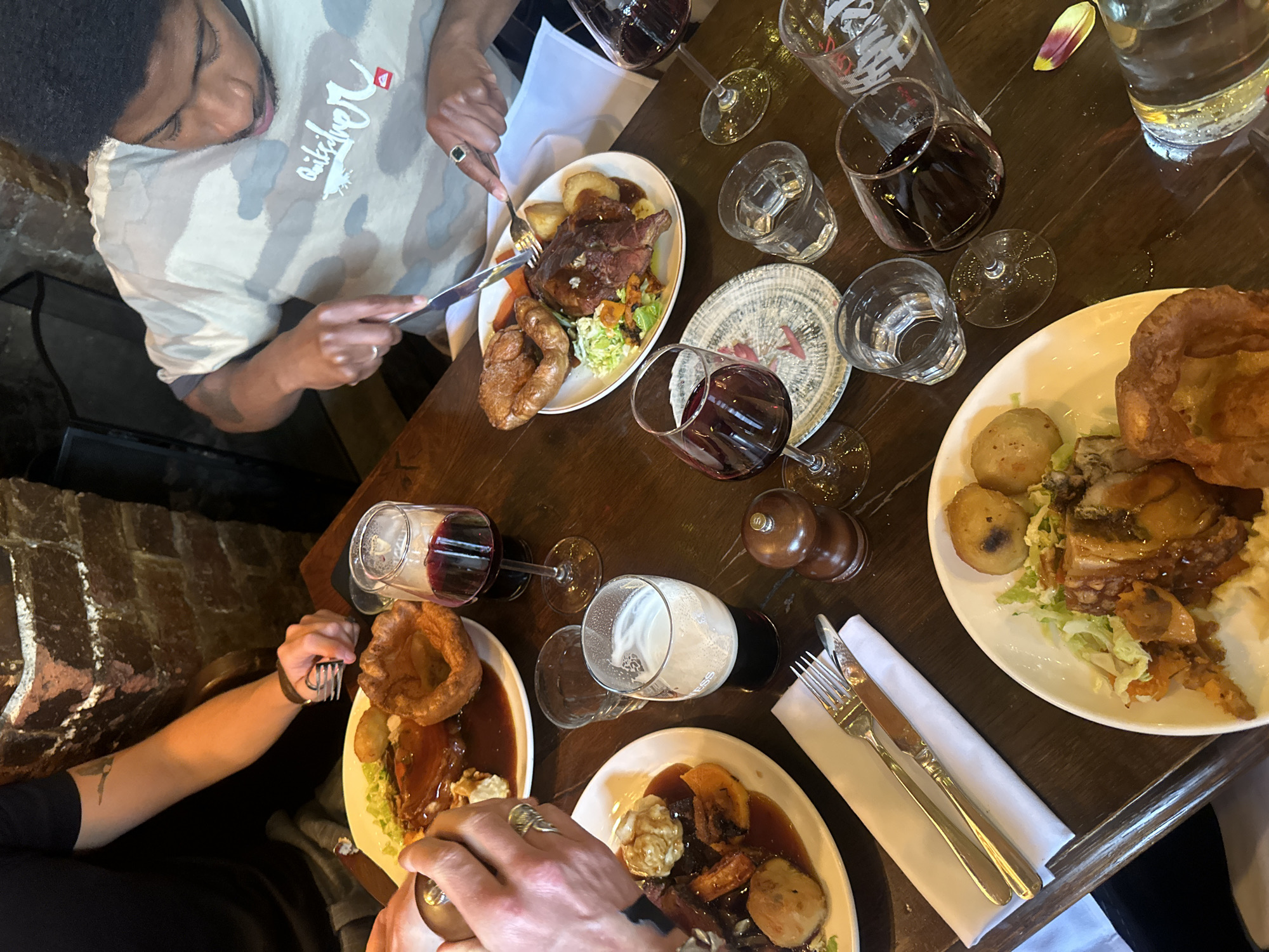 The image shows a table at a restaurant in London, United Kingdom, set with plates of traditional British Sunday roast dinners. Four people are partially visible around the table, two are using cutlery to eat. The dishes on the table consist of roasted meat, Yorkshire puddings, roast potatoes, and assorted vegetables. Several glasses containing red wine and beer are also present. Cutlery and napkins are neatly arranged. The table is dark wood.