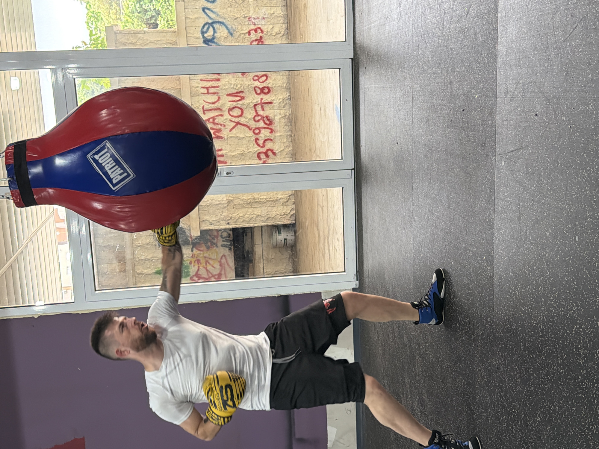 A man wearing a white t-shirt, black shorts, blue and black boxing shoes, and yellow boxing gloves is punching a red, white, and blue speed bag in what appears to be a gym. The gym has a purple wall and a gray tiled floor. A window is in the background with graffiti written on the glass. Based on the graffiti on the window, the location is Blagoevgrad, Bulgaria.