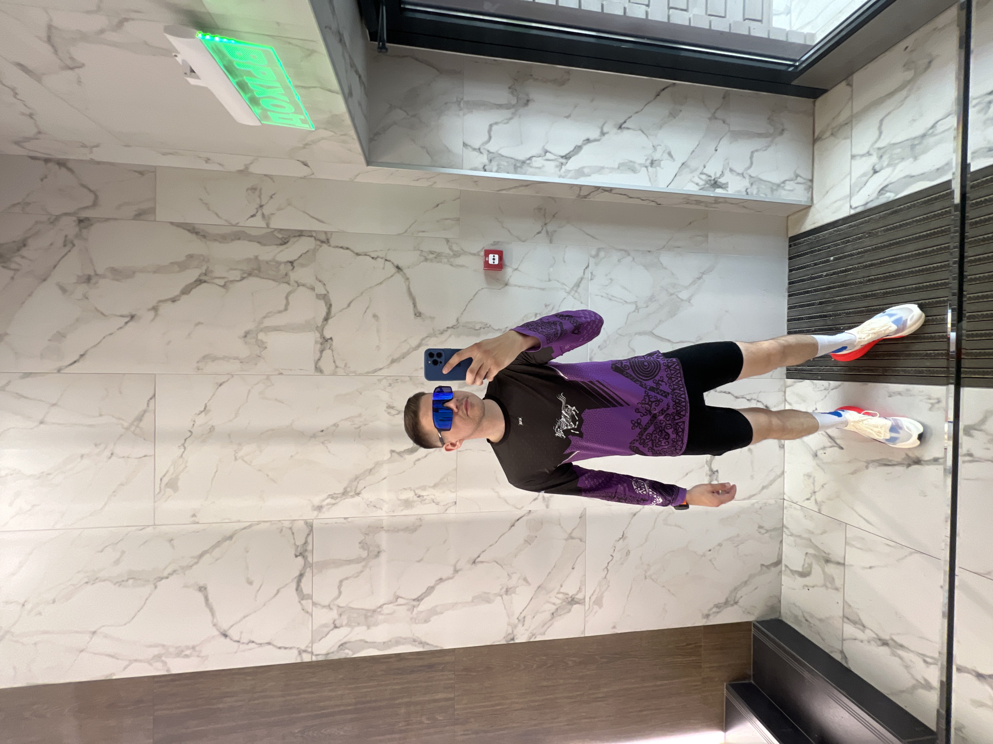 A man wearing blue sunglasses, a purple and black long-sleeved shirt, and athletic shoes is taking a selfie in a space with marble-patterned walls and floors. A sign is above him on the wall. The location is in Kazan, Russia.