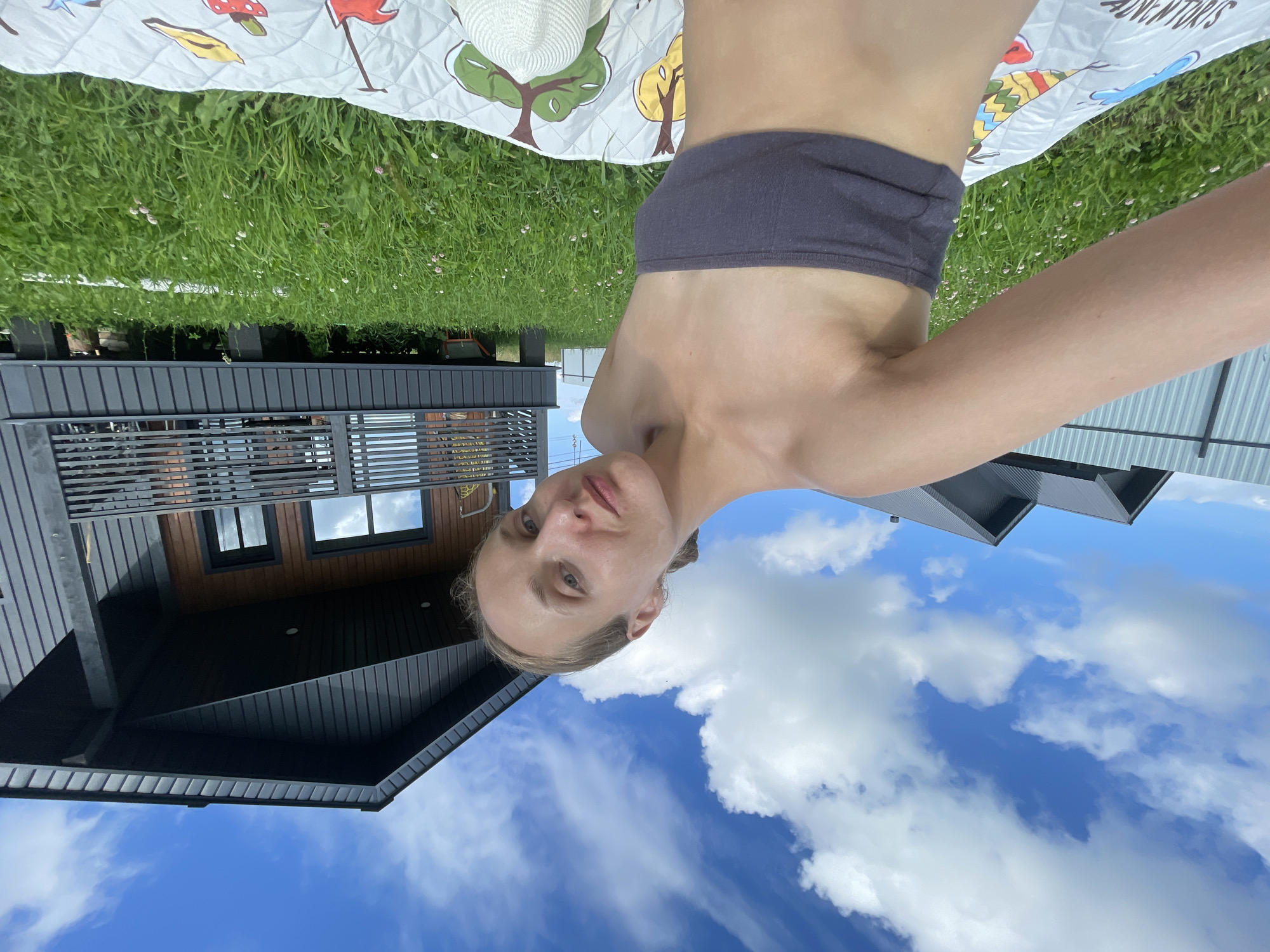 In the village of Podshivalovo, Russia, on a sunny day with a blue sky and scattered white clouds, a woman is taking a selfie outdoors. She is sitting on a colorful blanket spread across a lush green lawn dotted with small white and pink flowers. Behind her, a modern, dark-colored house with a wooden accent wall and a balcony stands on stilts. The house has dark-framed windows and a railing on the balcony. To the left of the house, another similar-style house is partially visible behind a metal fence. The woman is wearing a dark grey bandeau top, exposing her shoulders and upper torso. Her hair is pulled back into a bun. A white sun hat rests on the blanket near her. The overall scene depicts a relaxed, outdoor setting in a rural environment.