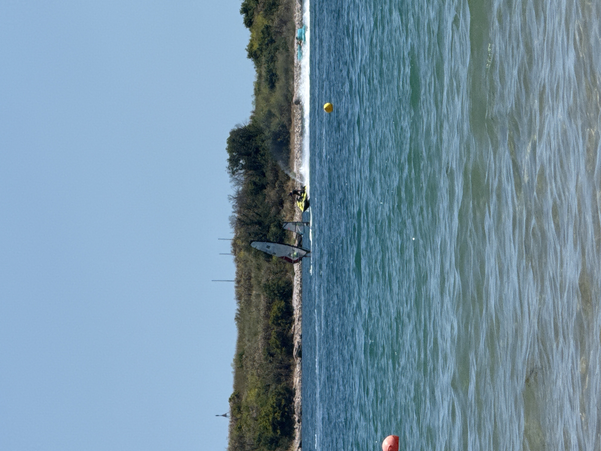 The image depicts a sunny day on the coast of Sozopol, Bulgaria, with a clear blue sky and calm, blue water. In the foreground, a red buoy is partially visible on the left side. The mid-ground shows several watercraft activities. Two windsurfers are sailing, one with a gray and white sail and the other with a smaller sail further back. A person is riding a yellow jet ski, creating a white spray of water. To the right of the jet ski, a turquoise boat with at least two occupants is also moving across the water, leaving a wake. In the background, a rocky shoreline is covered with dense green vegetation and trees. A thin black pole, likely a marker or part of a structure, is visible on the left side of the shore. Several other thin, vertical poles are visible further along the coastline. A yellow spherical buoy is floating in the water between the jet ski and the turquoise boat.