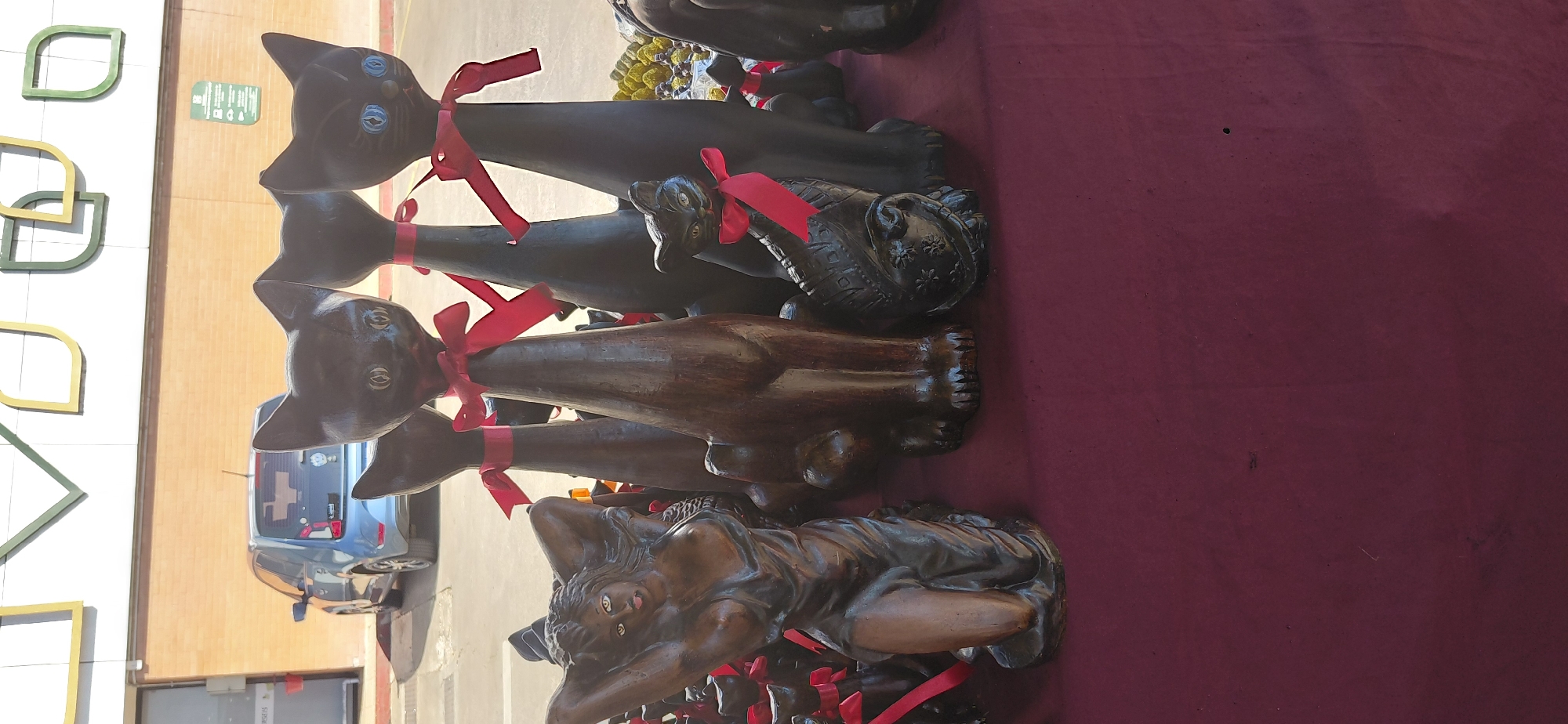 A sidewalk display of cat figurines and a human-shaped sculpture is shown in front of a building. The cat figurines are black with red bows around their necks. One of the cat figurines is smaller and appears to be holding a basket. The human-shaped sculpture is a dark-skinned female figure in a kneeling pose, with her hands behind her head and her tongue sticking out. The display is set up on a red table cloth. In the background, a blue car is parked in front of a brick building with a white and green sign. This scene appears to be located in Encarnación, Paraguay.