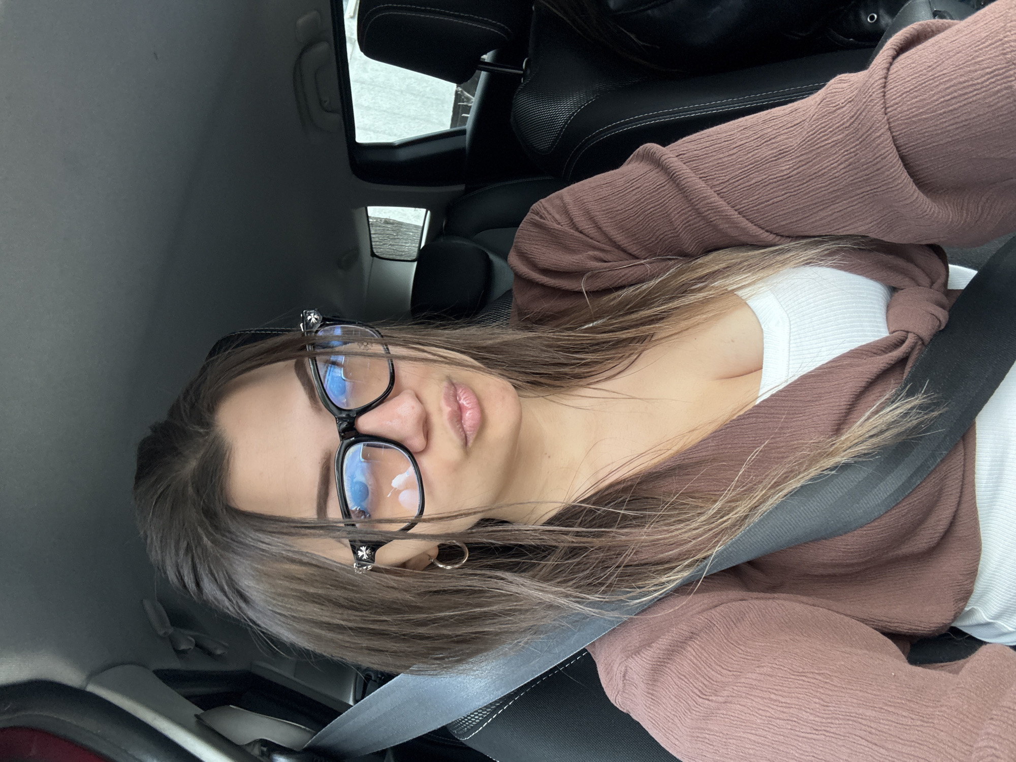 A woman with light brown hair and black glasses is in the driver's seat of a car, wearing a brown cardigan over a white top and a seatbelt. The interior of the car is visible, with black leather seats. Outside the car, blurred shapes suggest a city environment. It is not possible to determine if the scene is in Sofia, Bulgaria from the provided image.