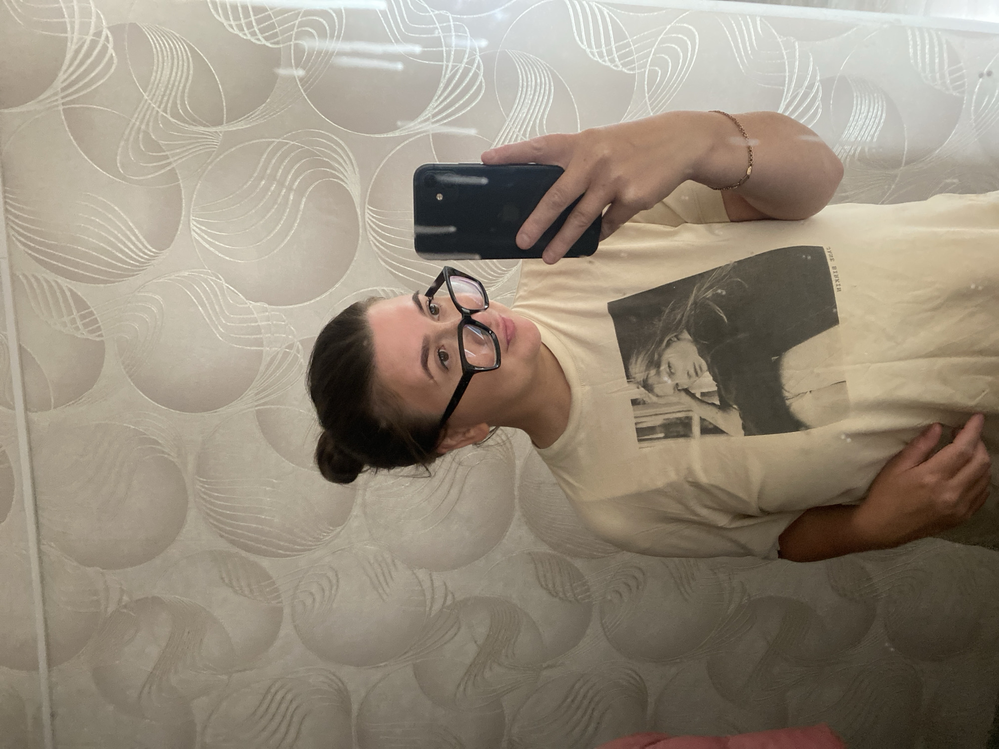 A woman with dark hair pulled up in a bun and wearing large-framed glasses is taking a selfie in a bathroom. She is wearing a light-colored t-shirt with a black and white image of a woman on it, and a thin gold bracelet on her right arm. She is holding a dark-colored smartphone in her right hand, with her thumb on the screen. The background consists of textured wallpaper with circular patterns in muted tones. The scene appears to be indoors, likely in a residential bathroom, and is not specific to any location within the city of Otchor, Russia.
