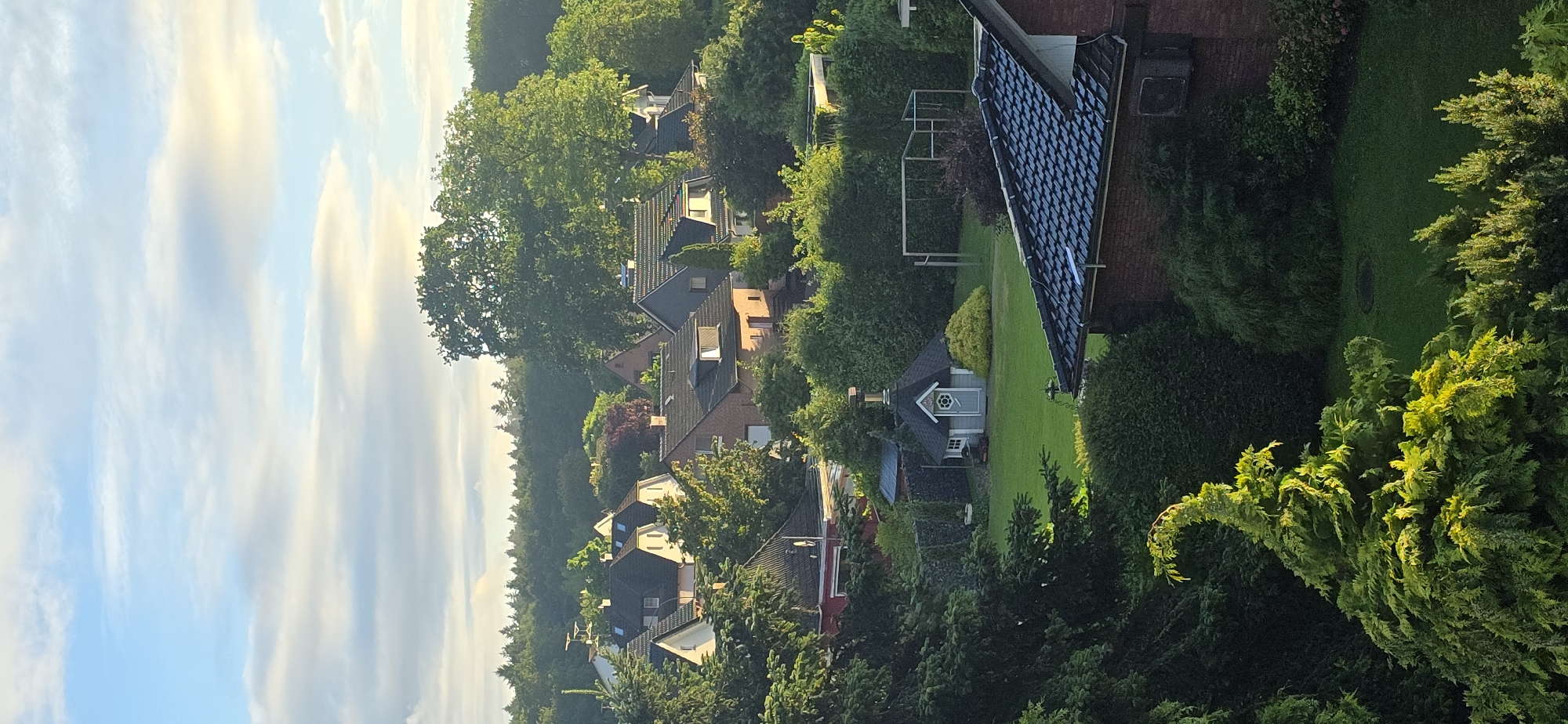 The image depicts a suburban residential area in Seevetal, Germany, with a view of houses nestled among lush greenery. The sky is partly cloudy, with hints of yellow and orange light suggesting either sunrise or sunset.
In the foreground, a vibrant green, conifer tree with upward-sweeping branches is illuminated by the sun, creating a dramatic silhouette. Beyond this tree, several houses with dark tiled roofs are visible, interspersed with dense foliage. One house in the mid-ground, constructed of red brick, has a dark tiled roof and an air conditioning unit mounted on its exterior wall.
Further into the scene, a quaint, hexagonal garden house with a dark roof, a chimney, and a decorative circular window is situated on a well-maintained green lawn. This structure is surrounded by manicured hedges and a yellow, rounded shrub. Adjacent to the garden house, a frame, possibly for a swing set or sports equipment, stands on the grass.
The background is dominated by a dense forest of mature trees, forming a natural boundary to the residential area. The overall impression is one of a tranquil, verdant, and well-established neighborhood. No people are visible in the image, and no specific actions or interactions are depicted.