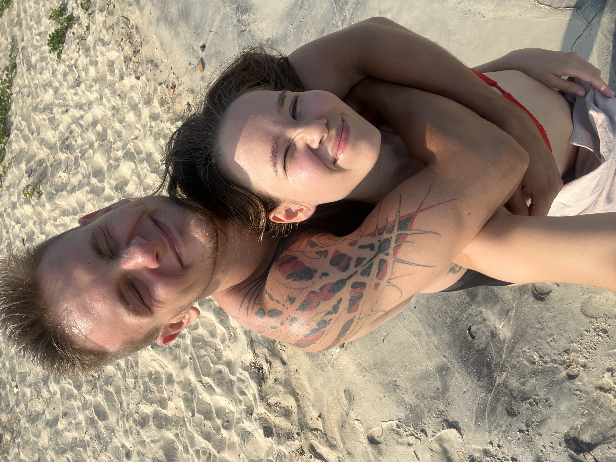 Two individuals, a man and a woman, are captured in a close-up, high-angle shot, lying together on a sandy surface, likely a beach in Nam Nha Trang, Vietnam. The woman is positioned on her back, her head turned towards the camera, her eyes squinted into a pleasant smile, suggesting enjoyment of bright sunshine. Her long, dark hair spreads around her head. Her arms are wrapped around the man, creating an intimate embrace. She wears a red bikini top, and a light-colored towel or garment is partially visible near her lower back.
The man lies next to her, facing the camera with his eyes closed or squinted, also appearing relaxed. He has short, light-colored hair and some facial stubble. A prominent tattoo featuring black and red abstract or tribal patterns covers his upper arm and shoulder. Both individuals appear to be shirtless or wearing minimal beachwear, and their skin shows the effects of strong sunlight with bright highlights and some shadows.
The setting is a sun-drenched beach, with fine, light-colored sand dominating the background. Patches of green vegetation are visible in the upper left corner, while some darker, possibly damp, sand areas appear towards the bottom right. The strong sunlight and the subjects' relaxed, smiling expressions convey a joyful and comfortable atmosphere. There is no visible text in the image.