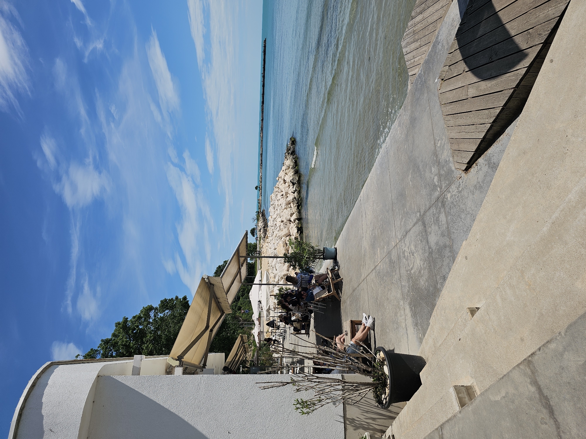 Here is a detailed description of the image:
**Scene Description:**
The image captures a sunny outdoor scene, likely a restaurant or cafe setting situated by the sea. The location appears to be on a raised concrete platform, transitioning to a lower level with concrete steps. The sky is blue with scattered clouds.
**Environment:**
* **Ocean:** A body of water is visible, likely the sea. A breakwater or pier extends into the ocean in the background.
* **Vegetation:** There are trees and plants in the background and in large pots near the seating area.
* **Structures:** A white building is present at the edge of the scene. Awnings provide shade over the seating area.
**People:**
* People are sitting at tables under the awnings. There seem to be several small groups dining or conversing.
* A person is reclining on a lounge chair at the edge of the concrete platform. Their legs and shoes are visible.
**Objects:**
* Tables and chairs are arranged under the awnings.
* Large plant pots contain trees or shrubs.
* A concrete breakwater extends into the sea.
* A wooden planked area is on the right side of the concrete platform.
**Actions/Interactions:**
* People appear to be dining or socializing at the cafe.
* The person on the lounge chair seems to be relaxing.
**Location:**
* City: Unknown
* Unknown.
**Overall Impression:**
The image depicts a leisurely scene, likely during the daytime, at a seaside cafe or restaurant. It suggests a warm climate.