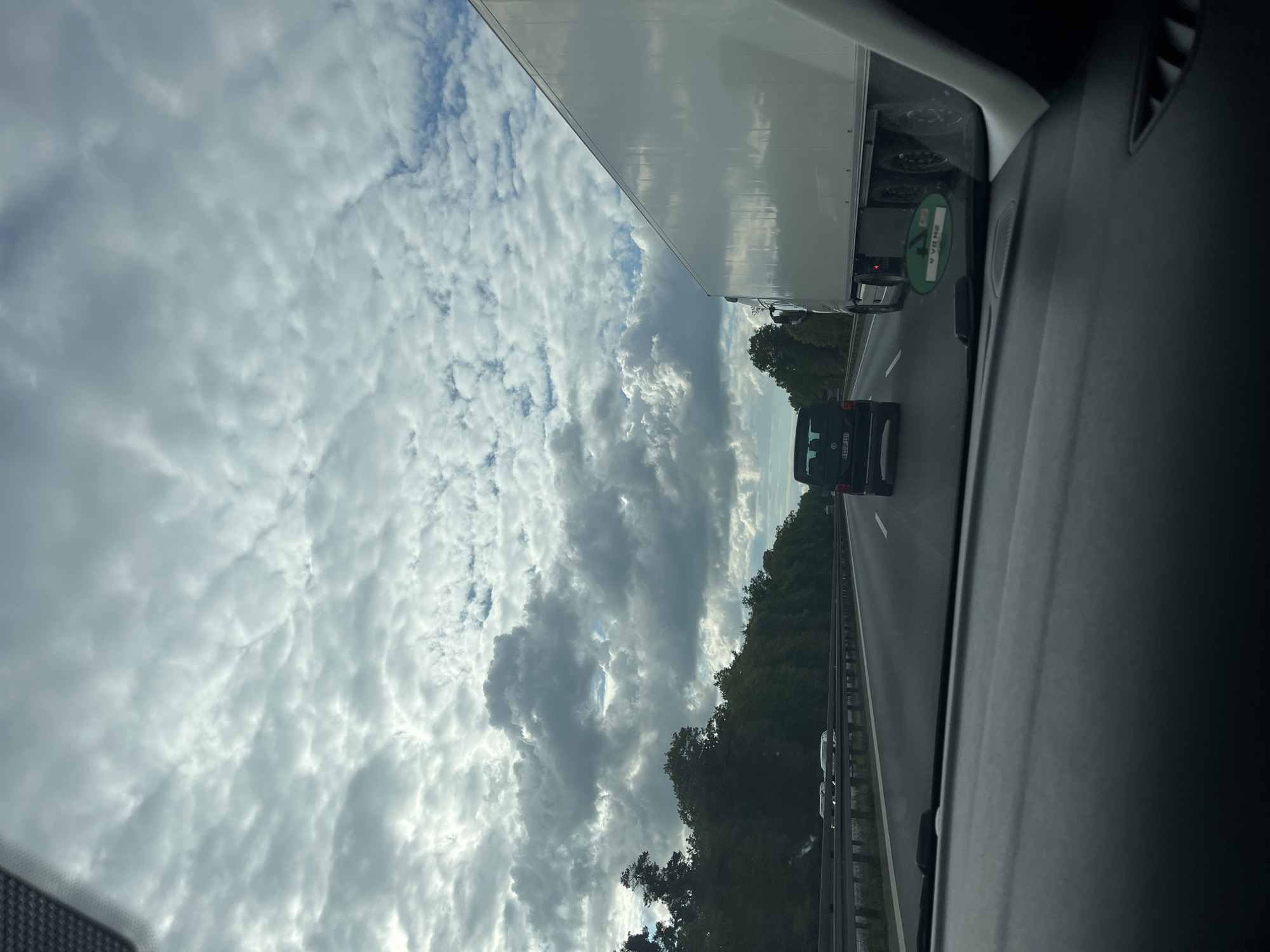 The image shows a view from inside a car on a highway. To the left is a large white truck, and ahead are several other vehicles. Trees line the right side of the road. The sky is overcast with numerous clouds. The location is Unknown, Unknown.