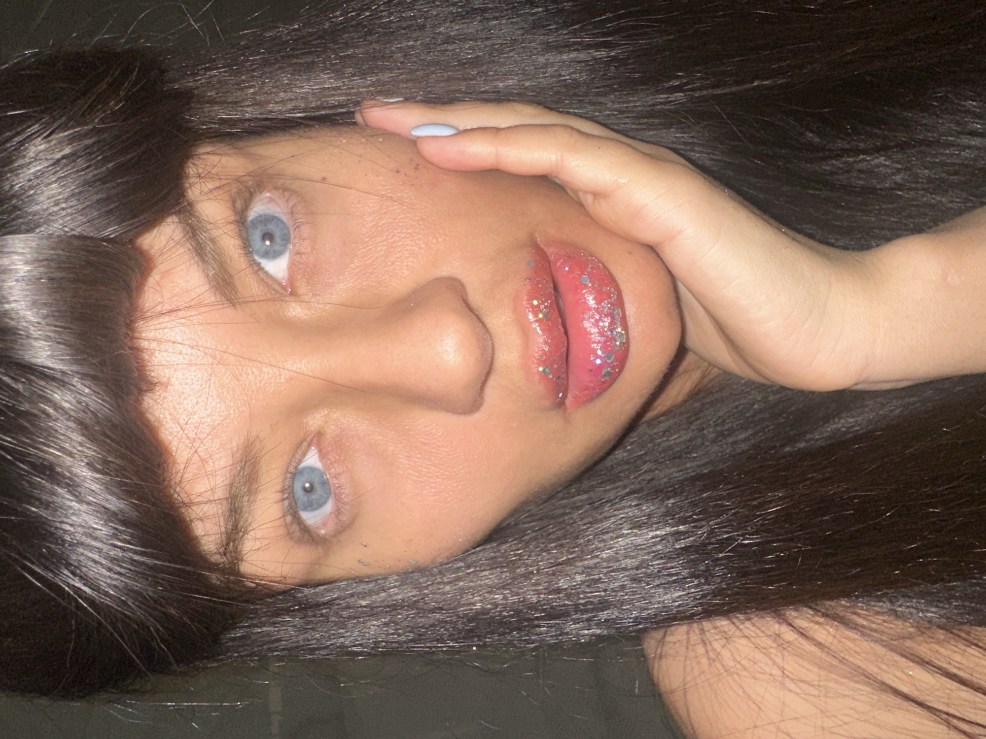 A close-up, vertically oriented portrait features a person with fair skin, striking blue eyes, and long, dark hair. The subject’s head is tilted, as if lying down or posing at an angle. Dark, straight hair frames the face, with some strands obscuring parts of the forehead and cheeks. The person gazes directly forward with an intense, yet serene expression.
Notable makeup includes a glossy, glittery pink lip color, with scattered specks of silver glitter visible on the cheeks, forehead, and near the eyes, suggesting an artistic or celebratory style. A hand rests gently on the upper right side of the face, near the temple and forehead, with the thumb touching the cheek. One visible fingernail on the index finger is painted a pale light blue. The background is dark and indistinct, indicating an indoor setting or low-light conditions. There is no visible text or explicit location cues within the image. The overall impression is a contemplative or artistic portrait.