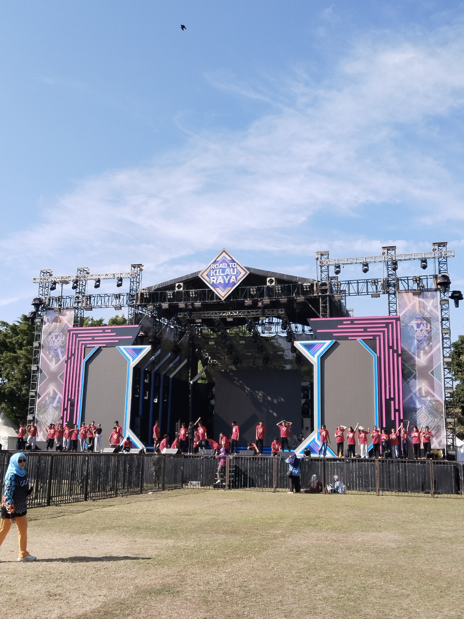 This is a medium-shot, eye-level, full-color photograph of an outdoor stage setup with a crowd of people in front of it. The stage is large and elaborate, with a backdrop displaying a diamond-shaped logo that reads "ROAD TO KILAU RAYA". The stage is lit with multiple spotlights and is supported by a complex metal truss system.
A group of performers, mostly young people dressed in red t-shirts, are on the stage, some in poses. A black metal fence separates the stage from a grassy area where the audience is gathered. In the foreground, a woman wearing a blue headscarf and orange pants walks across the grass, casting a shadow. The sky above is bright blue with scattered white clouds, and a single black bird is visible in flight in the upper left quadrant of the image. The overall impression is that of a daytime outdoor event, possibly a concert or festival, taking place in a public park or open field within Kecamatan Pati, Indonesia.