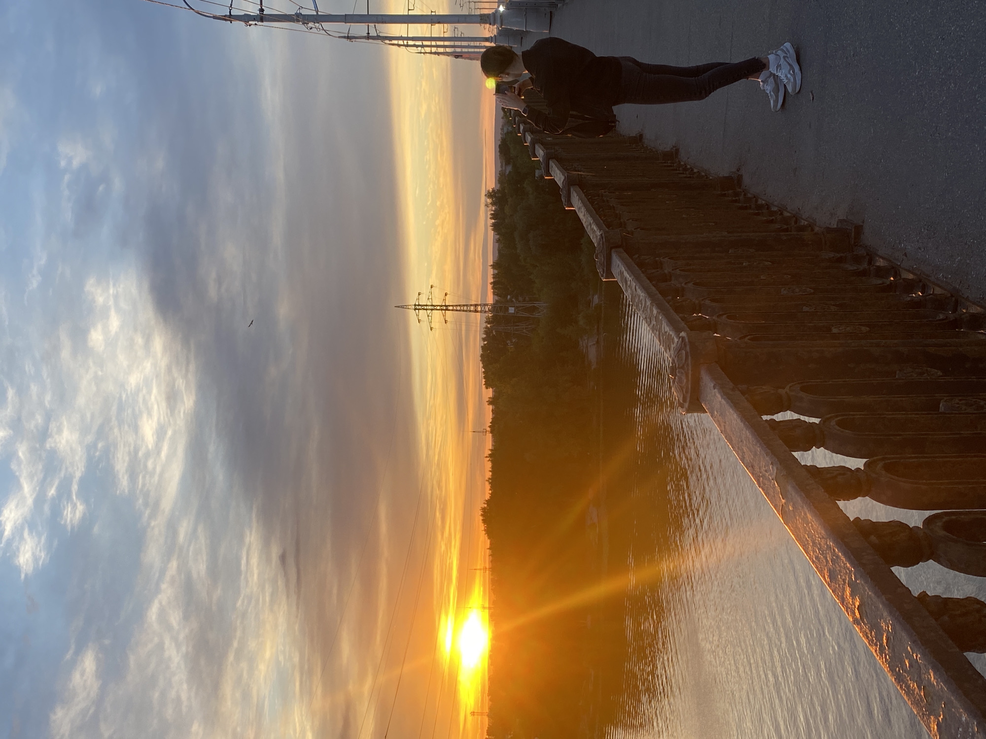 The media file depicts a person standing on a bridge overlooking a body of water at sunset. The sky is filled with hues of orange, yellow, and blue, indicating the sun is low on the horizon. Wispy clouds are scattered across the sky, catching the warm light of the setting sun. A person, dressed in dark clothing, is leaning on a decorative metal railing, appearing to be looking at their phone. White sneakers are visible on their feet. The bridge has an old-fashioned metal railing with intricate designs. Below the bridge, the water reflects the colors of the sunset, creating a shimmering effect. Trees line the opposite bank of the water, and in the distance, power lines and towers are visible against the sunset. The scene evokes a sense of peaceful contemplation as the day comes to a close. The time of day is sunset, and the weather appears clear with scattered clouds. The presence of power lines and the architectural style of the railing might suggest an urban or semi-urban environment. There is no visible text in the media file.