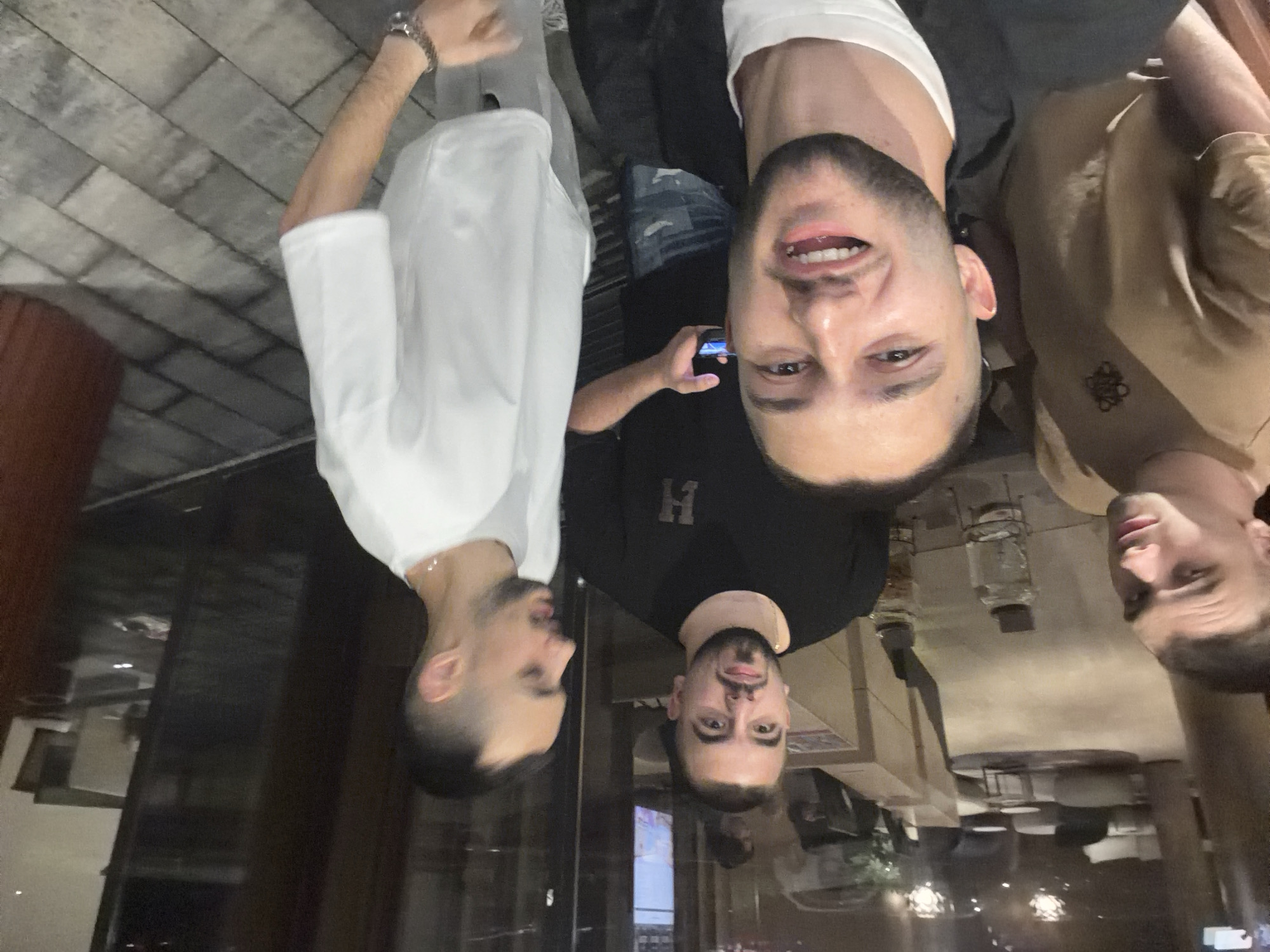 This indoor photograph, likely taken in Sofia, Bulgaria, features four men in what appears to be a restaurant, bar, or lounge during the evening or night. The image is captured from a slightly upward angle, with a reflective surface in the foreground mirroring the subjects and the dimly lit background.
Three men are clearly visible, while a fourth is partially obscured. In the center of the frame, a man with a short beard and dark hair smiles broadly, looking directly at the camera. He is wearing a white t-shirt. To his left, another man with a beard and short hair is visible from the chest up, also wearing a white t-shirt and a watch on his left wrist. He appears to be holding a glass or bottle. To the right of the central man, a third man with a beard, wearing a beige shirt, is partially visible, looking slightly away from the camera. His shirt features a dark, stylized, interlocking logo or emblem. Behind the central man, a fourth man, wearing a black t-shirt with a white uppercase 'H' printed on it, holds a mobile phone and is partly obscured by the central figure.
The setting includes a tiled or brick-patterned wall section in the background, along with tables and ambient lighting, all contributing to a relaxed, social atmosphere. The men's expressions suggest a convivial mood.