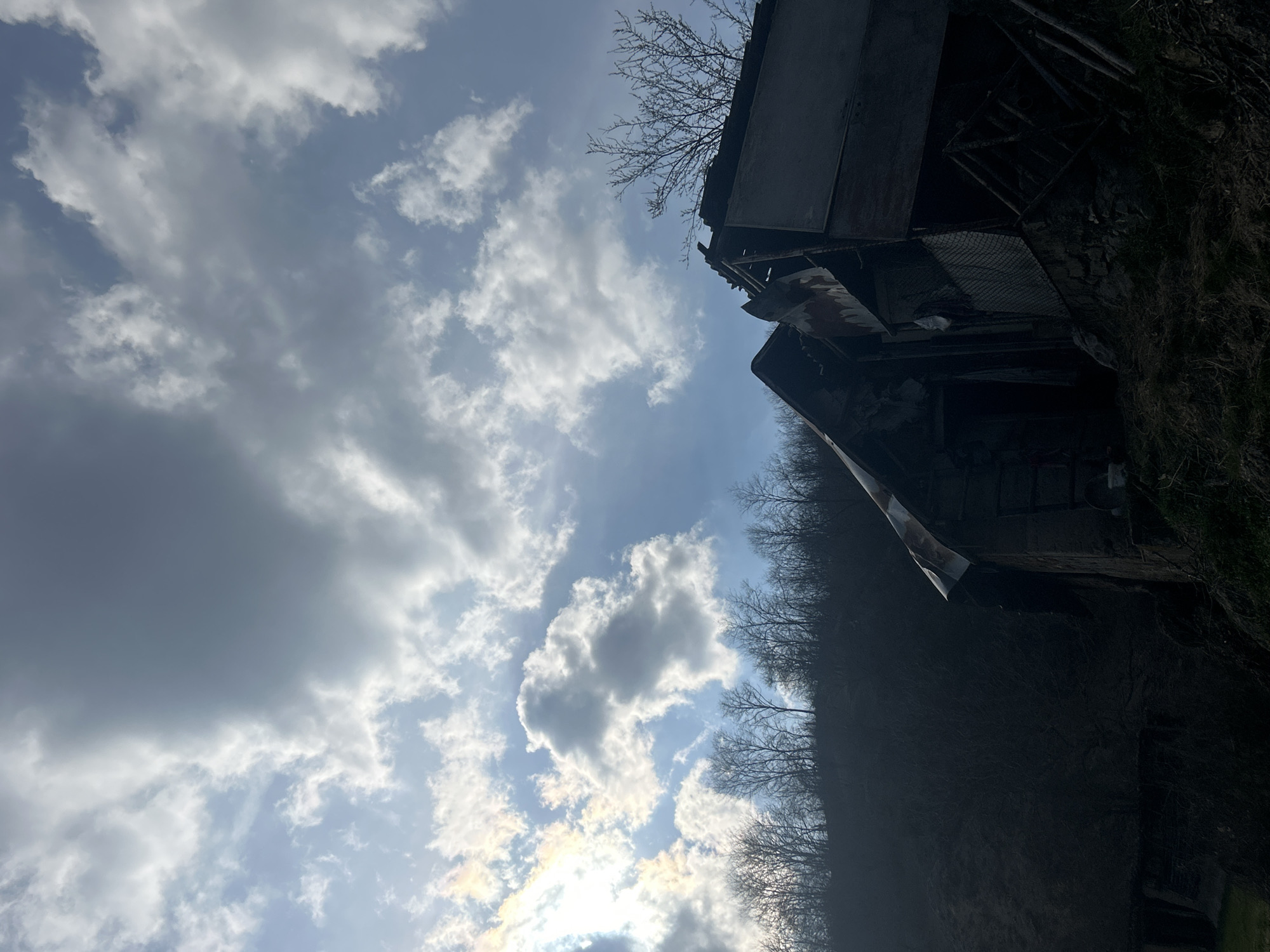 The scene depicts a severely dilapidated building structure situated on a hillside in Zlatograd, Bulgaria. The structure, possibly an abandoned outbuilding, occupies the lower right portion of the frame and is rendered in deep silhouette against a bright, partially cloudy sky. Its roof is composed of heavily rusted, corrugated metal, exhibiting significant damage and partial collapse. The visible substructure includes what appears to be a stone foundation, with upper sections showing exposed wooden framing and a section of mesh or chain-link fencing. Directly behind the building and extending across the mid-ground is a steep, dark hillside densely covered with bare deciduous trees. The sky features a clear blue interspersed with white and grey cumulus clouds, with the sun prominently visible in the upper left, causing strong backlighting and glare. The immediate foreground consists of shadowed, overgrown terrain with dry grass and sparse vegetation. No human or animal subjects are present, nor are any activities depicted.