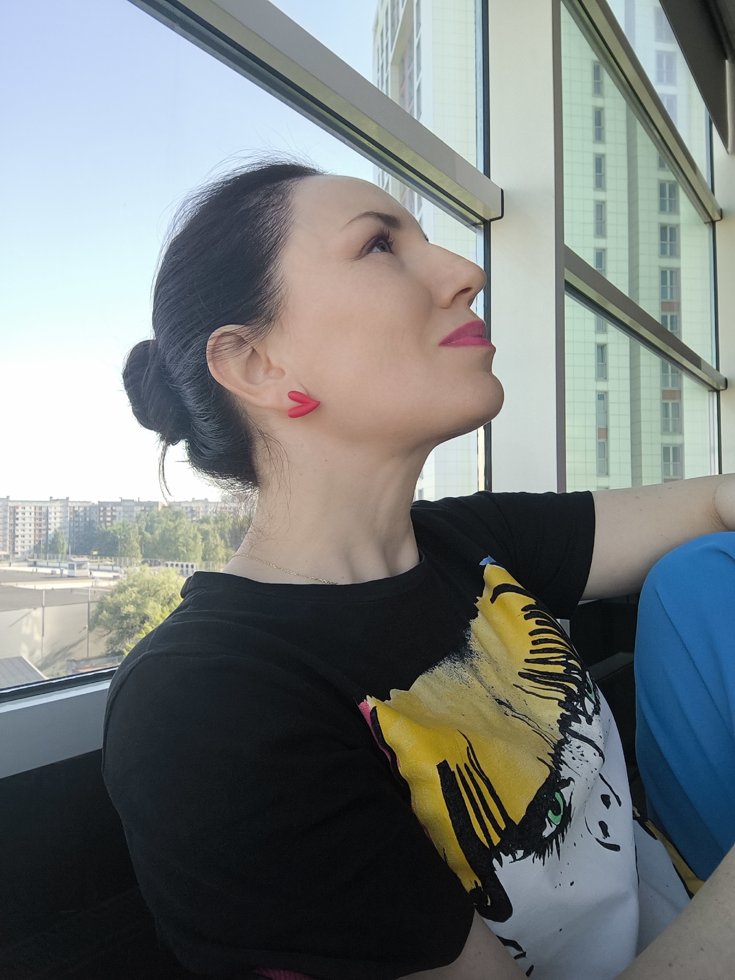 A woman is depicted from the chest up, positioned in profile as she looks upwards and towards the right. She has dark hair styled in a bun and is wearing bright pink lipstick and red, arrow-shaped stud earrings. A thin, gold-colored necklace chain is visible around her neck. She wears a black t-shirt featuring a graphic design of a face, with yellow and white elements and green eyes, partially visible on her chest. To her right, a vibrant blue fabric can be seen, possibly part of her clothing.
The setting appears to be indoors, next to a large window. Through the window on the left, a bright, clear blue sky is visible above a landscape of distant buildings and green trees, suggesting an urban or suburban environment. On the right side of the window, a tall, modern building with many rectangular windows and a light-colored facade with some orange vertical accents is visible, reflecting the bright sky. The window frame itself is prominent, with modern, metallic-looking dividers. The time of day appears to be sunny daytime, with clear weather. No visible text is present in the image.
