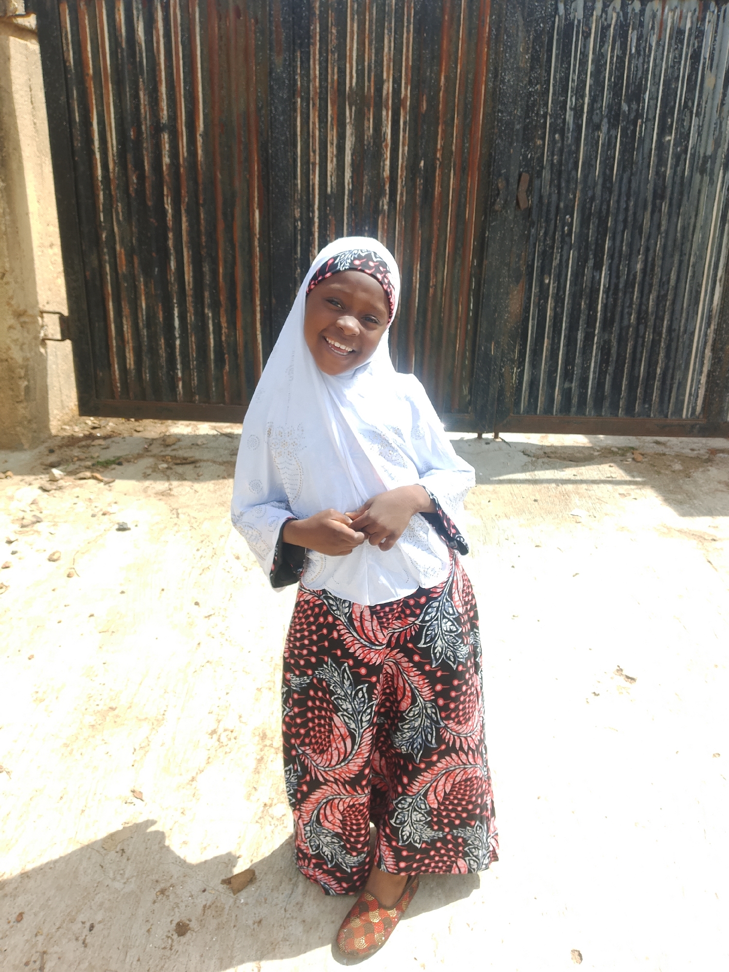 A young girl wearing a white hijab and a light blue patterned top with loose-fitting, black and red patterned pants stands facing forward, smiling. Her hands are clasped together in front of her. She is wearing patterned shoes. The background consists of a rusted corrugated metal fence and a light-colored, rough ground surface. The scene appears to be outdoors, likely in Jos, Nigeria, given the context. The lighting suggests it is daytime.