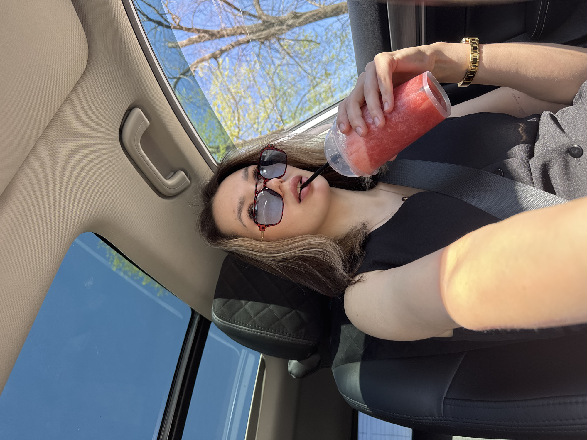 A woman wearing sunglasses and a gold bracelet is drinking from a clear plastic cup with a straw, inside a car. She has a black shirt and grey pants. She has a seatbelt on. The sky and tree branches are visible through the car window. The location is unknown.