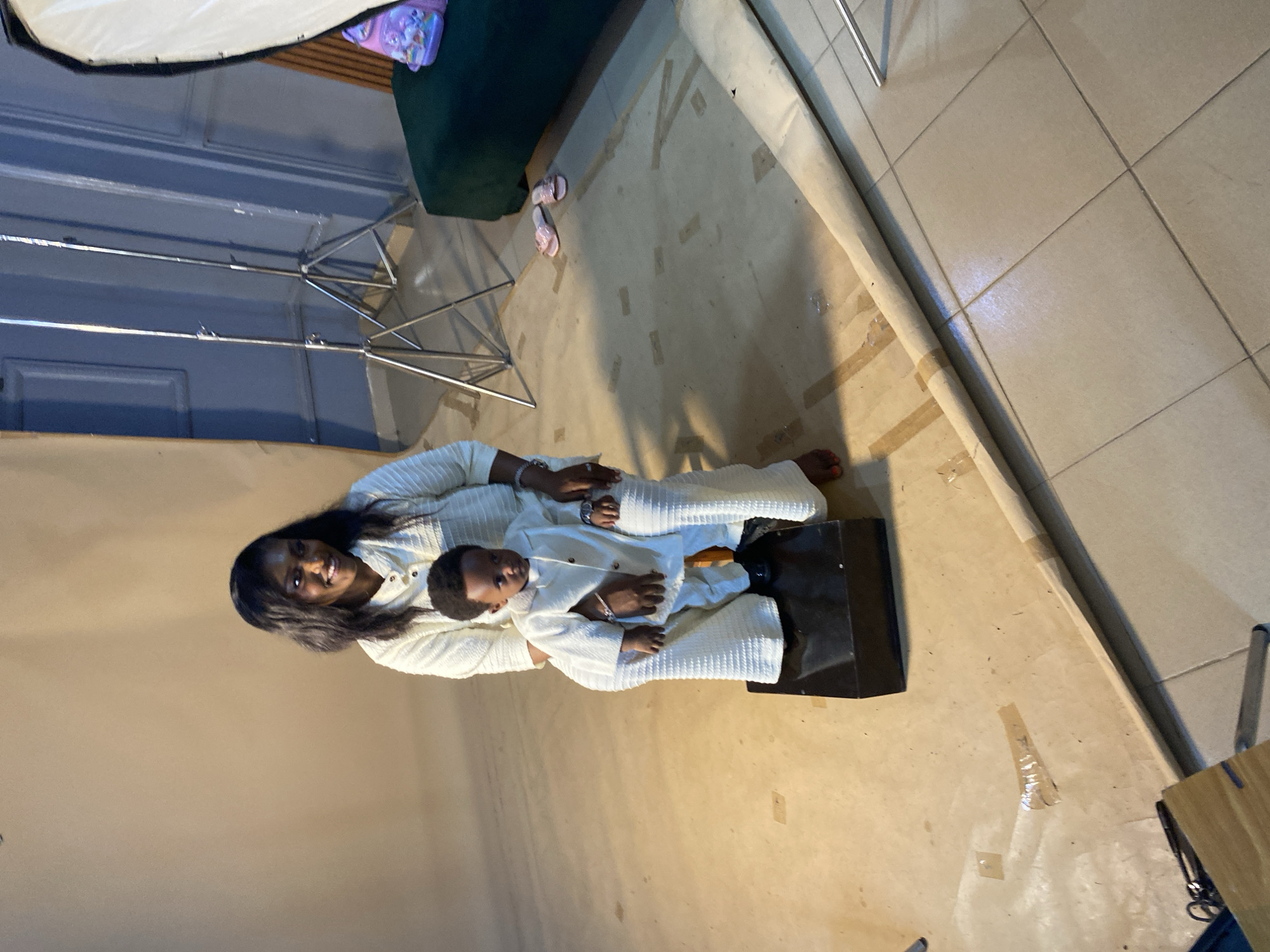In a studio setting, a Black woman with long dark hair is seated, holding a young Black child on her lap. Both are wearing matching cream-colored outfits. The woman is smiling and looking to her right. The child is looking forward. They are positioned in front of a light brown backdrop. To the right of the woman and child, there are two professional studio lighting stands, a portion of a dark green couch with a pink bag on it, and a large softbox light diffuser. The floor is tiled, with a large roll of brown paper unfurled across it. Scattered on the floor are pink flip-flops. The overall ambiance suggests a professional photoshoot. The scene is located in Bukuru, Nigeria.