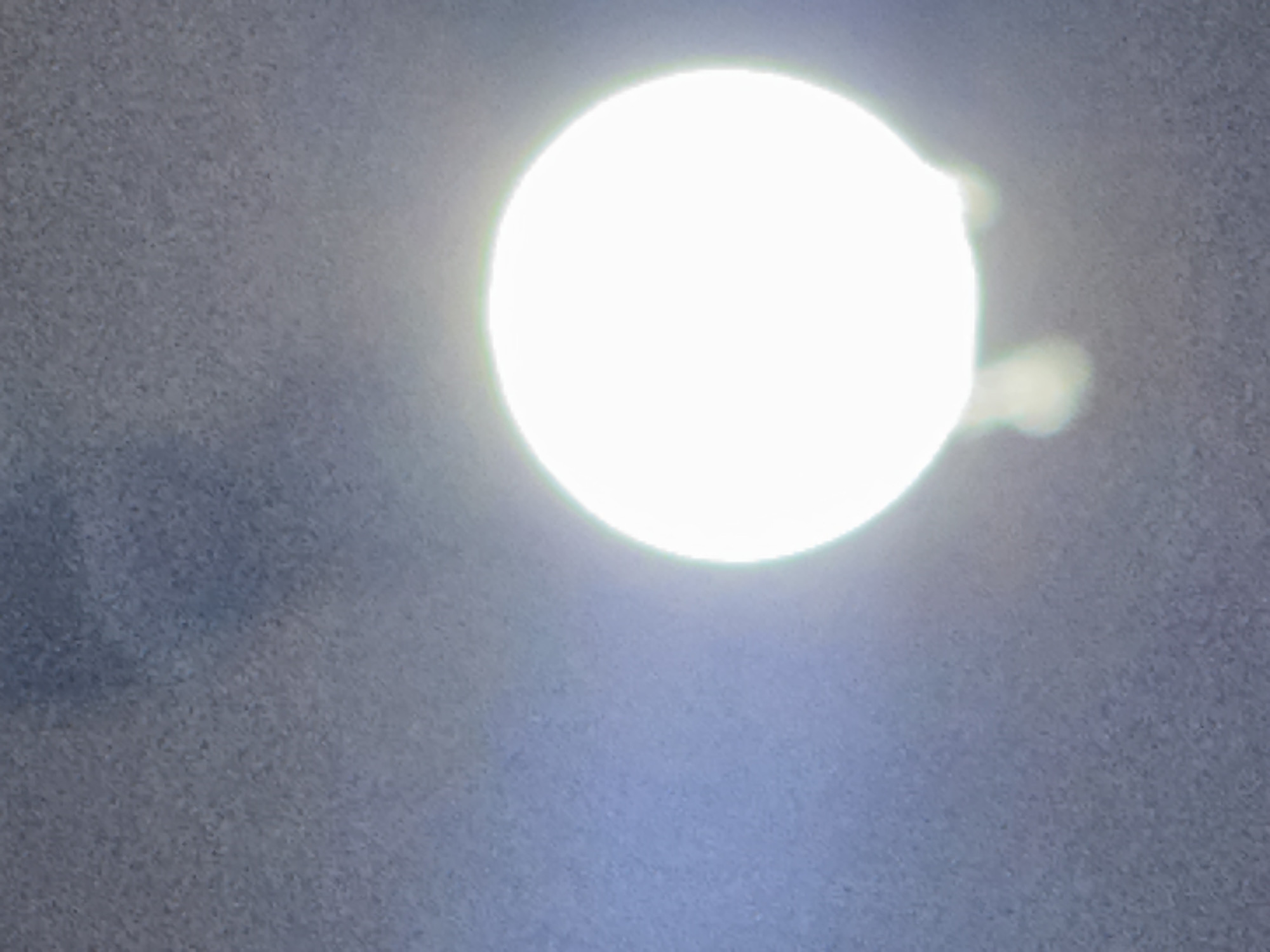 The image shows a brightly lit, out-of-focus circle against a grainy, grey background. There are hints of blue light emanating from the left side of the circle, suggesting a light source such as a lamp or a very bright bulb. Below the circle, there are blurry, wispy white shapes that appear to be a consequence of the light source. The overall scene lacks any discernible urban context and does not provide any information about the city of Navbahor, Uzbekistan, or any specific details of people, objects, or actions.