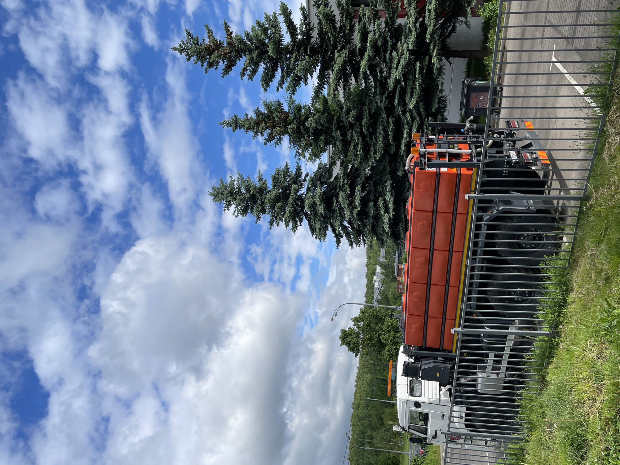 The image depicts a white and orange utility truck with multiple containers or tanks mounted on its chassis, parked beside a metal fence along a paved road. Evergreen trees stand near the truck, contrasting with the blue sky dotted with puffy white clouds. The location is in a wooded area, with greenery and buildings visible in the background. The scene takes place outdoors, under daytime lighting. Given the presence of a vehicle known to be in use in Moscow, Russia, the location is likely in the city of Moscow, Russia.