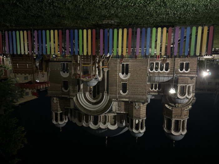 The image captures an inverted nighttime scene, primarily featuring a large, ornate building, likely a church, viewed from a slightly elevated position.
In the immediate foreground, running across what would be the bottom of the image if it were correctly oriented, is a colorful picket fence. Each picket is painted a different bright color, creating a vibrant sequence of red, orange, yellow, green, blue, purple, and various shades in between. Beyond this fence, there is dark foliage, possibly grass or bushes, indicating a park or green area.
The main subject is a substantial, multi-domed building, characteristic of Orthodox architecture, constructed from light brown brickwork. It features multiple arched windows, many with intricate white frames or decorative elements. The building has several domes of varying sizes, some topped with crosses, and intricate architectural details such as cornices, moldings, and semi-circular porticoes. A large, grand entrance or facade with a prominent arched structure is visible towards the center. On the right side of the building, a hanging lamp fixture casts light onto the brick facade.
To the left of the main building, another structure with a reddish roof and simpler windows can be seen, partially obscured by dark foliage. The overall setting is dark, indicating it is nighttime, with artificial lights illuminating parts of the main building and a bright light source visible further in the background on the right. The ground or surface below the building is very dark, possibly water reflecting the distant lights or simply a dark, unlit area.
No text is visible in the image. The scene conveys a sense of quiet grandeur, with the brightly colored fence offering a contrast to the solemn architecture of the illuminated building at night.