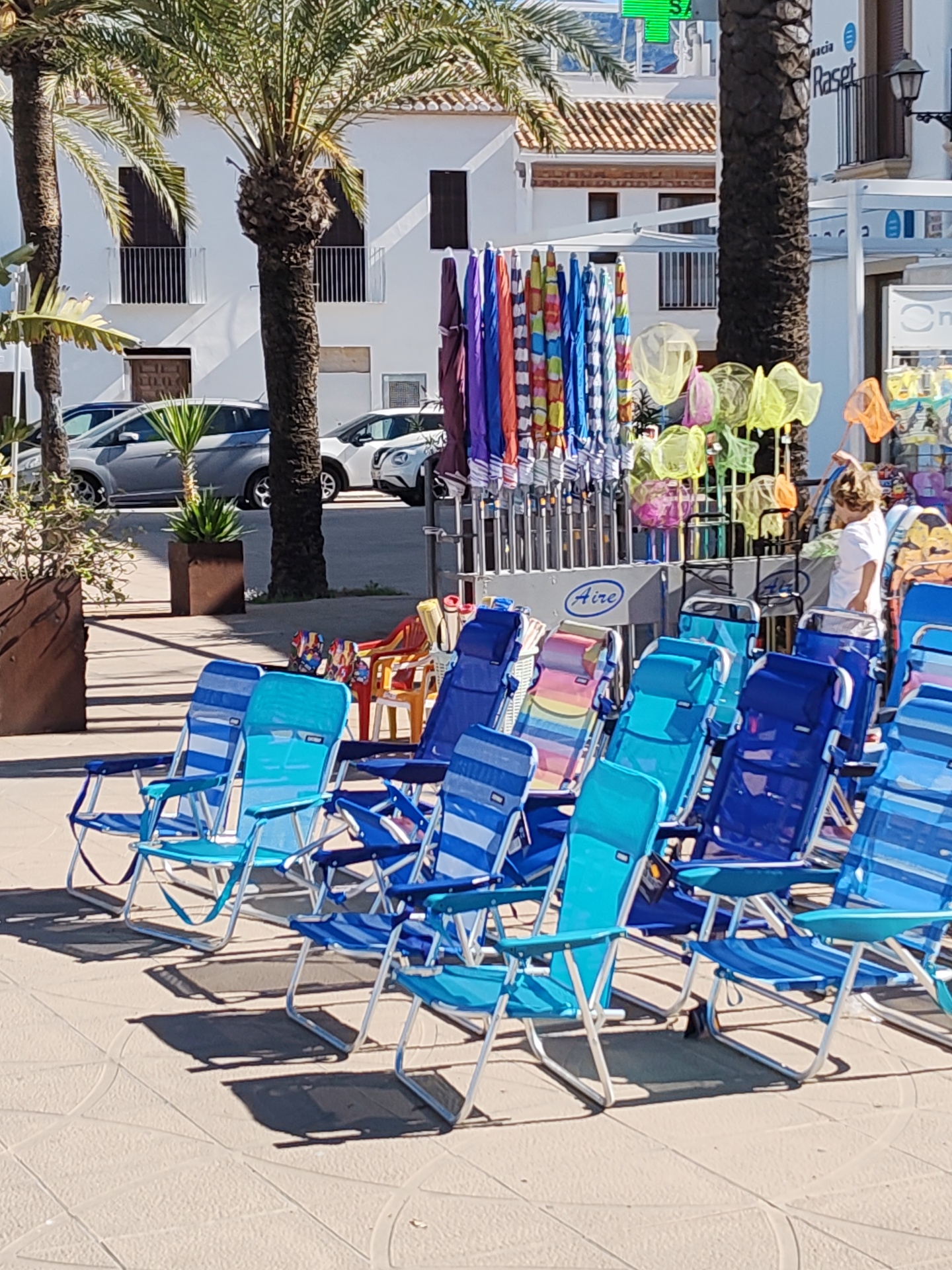 This outdoor scene depicts a retail display of beach supplies for sale in Dénia, Spain. The primary focus is on numerous folding beach chairs in shades of blue. Behind the chairs, there's a stand holding rows of closed, colorful beach umbrellas and various colorful fishing nets. A woman wearing a white shirt is standing next to the stand of fishing nets and appears to be browsing through the merchandise. In the background, there are several parked cars and white buildings, some with palm trees in front of them. The merchandise stand has "Aire" printed on it. The scene is well-lit, suggesting a sunny day.