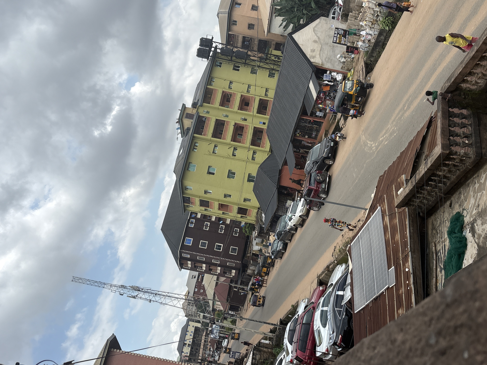 The image depicts a street scene in Nkpor, Nigeria, with a focus on a moderately busy road lined with buildings and parked vehicles. The sky is overcast with dramatic clouds. On the left side of the street, several parked cars are visible, including a silver sedan and a dark-colored SUV. Further down the street, a light pole stands tall. The buildings flanking the road vary in architectural style and color, with a prominent light green, multi-story apartment building on the right. Several shops with dark roofs are also present, displaying goods. The road itself is paved with asphalt and shows signs of wear. Various modes of transportation, including a yellow and black auto-rickshaw, are present on the road. Several individuals are walking on the sidewalks and crossing the street, engaged in daily activities. The overall impression is that of a typical urban street in a developing city, with a mix of residential and commercial activity.