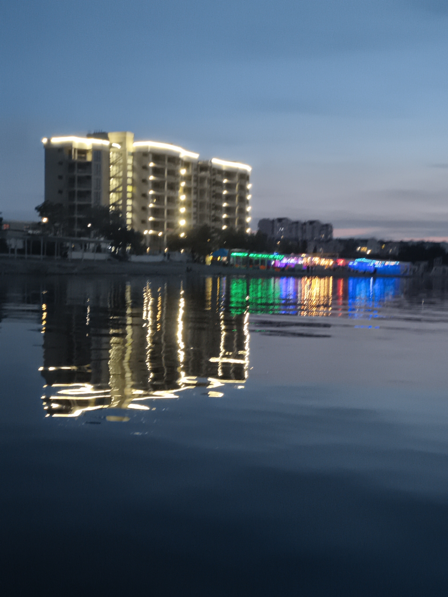 The image displays a nighttime or late dusk scene in Sevastopol, Ukraine, characterized by illuminated urban structures reflected in a body of water.
The foreground is dominated by a calm, dark expanse of water, likely a bay or large inlet. This water surface exhibits distinct reflections of the lights from the distant shore, appearing as vertically elongated, shimmering streaks of warm white, green, yellow, red, and blue.
In the midground, along the opposite bank of the water, several buildings are visible. On the left, a large, multi-story building or complex with a light-toned facade is prominently illuminated. It features extensive warm white strip lighting that traces its architectural lines, particularly along horizontal elements and potentially outlining the roofline. Numerous individual warm white lights are also visible within its structure, emanating from windows or interior spaces. This building appears to be a modern residential or commercial complex.
To the right of the large building, a cluster of lower-rise structures along the waterfront is illuminated with vibrant, multicolored lights. These include distinct sections of green, yellow, red, and bright blue lighting, suggesting the presence of various establishments such as restaurants, cafes, or recreational venues. These colorful lights are also clearly reflected in the water.
The background shows a twilight sky, transitioning from a deep blue at the top to a lighter blue-grey towards the horizon. Faint silhouettes of additional urban buildings and scattered distant lights are discernible further back, indicating an extended city environment. No individuals, specific actions, or interactions are clearly discernible within the frame.