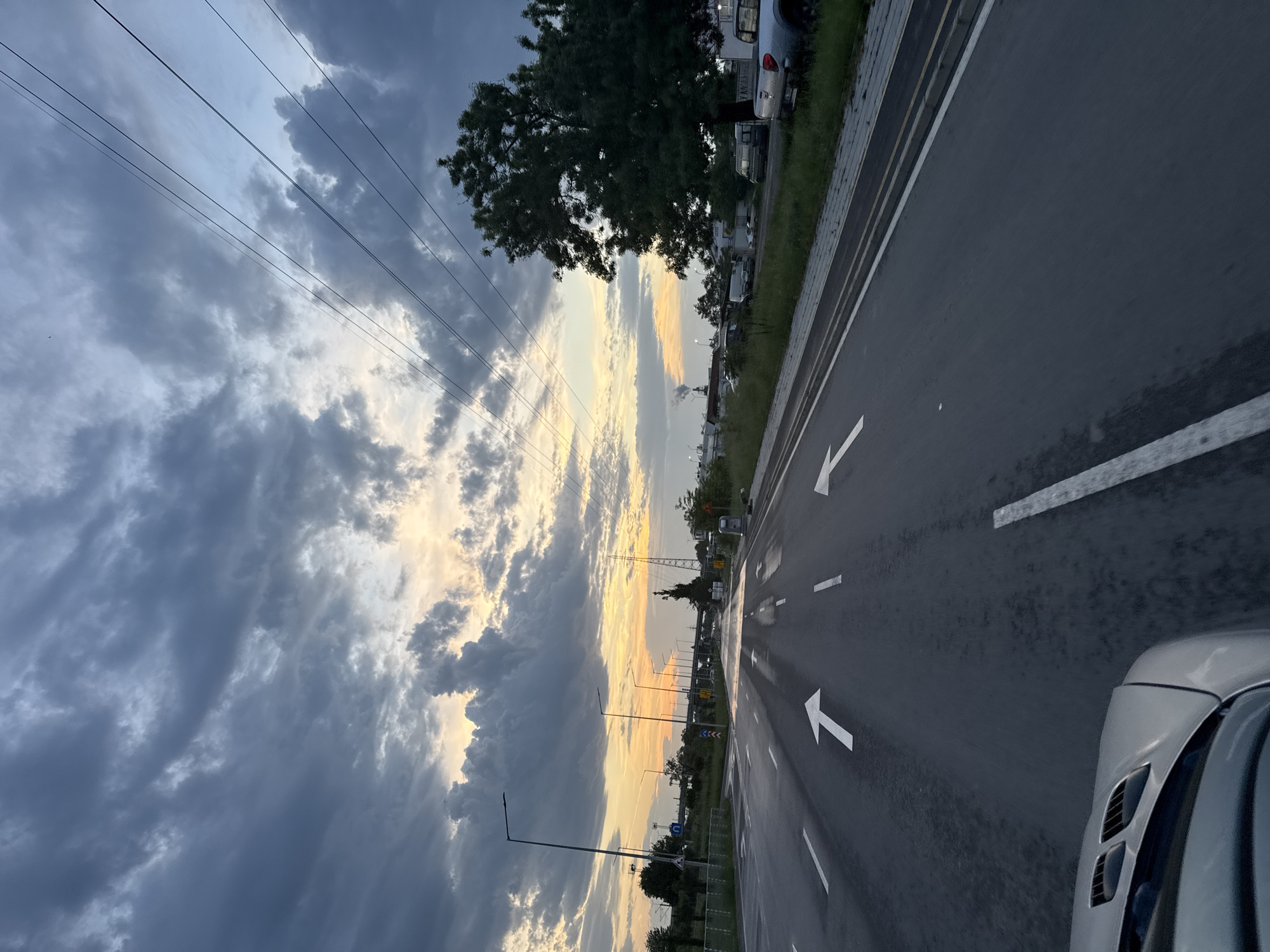 The image presents a wet road under a dramatic sky with partly cloudy conditions and a sunset glow. The location appears to be a multi-lane road with white directional arrows painted on the surface. Along the side of the road, there is a strip of grass and parked vehicles. Utility poles and wires are visible, extending towards the horizon. Part of a car is visible in the lower right corner. Although the location is in Burgas, Bulgaria, there is no available evidence to precisely identify the specific area within the city.