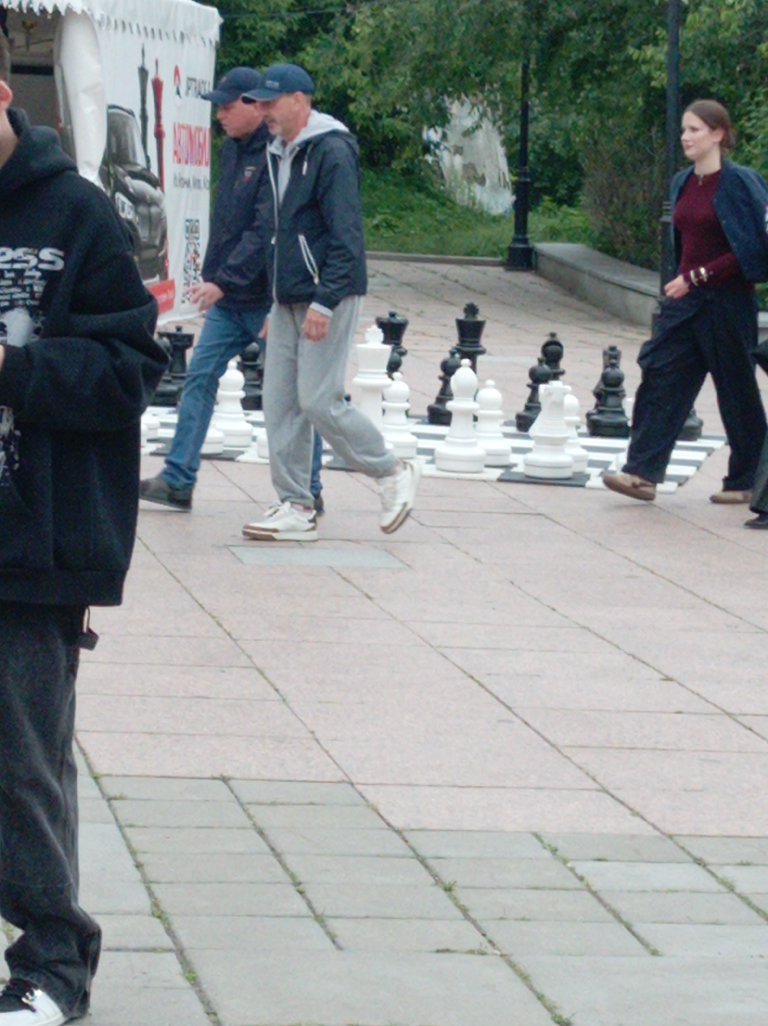 In Vladivostok, Russia, a group of people are engaged in a game of oversized chess on a paved outdoor area. In the foreground, a person wearing a black hooded sweatshirt and dark pants is partially visible, facing away from the camera. To their right, two men are standing on the chess board. One man, wearing a dark jacket and jeans, appears to be moving or observing a chess piece. The other man, in a grey hooded jacket and light-colored pants, also seems involved in the game. Further to the right, a woman in a dark jacket over a maroon top and dark pants is walking. The environment suggests a public space with a tent-like structure in the background and trees beyond. The chess pieces are large and predominantly black and white.