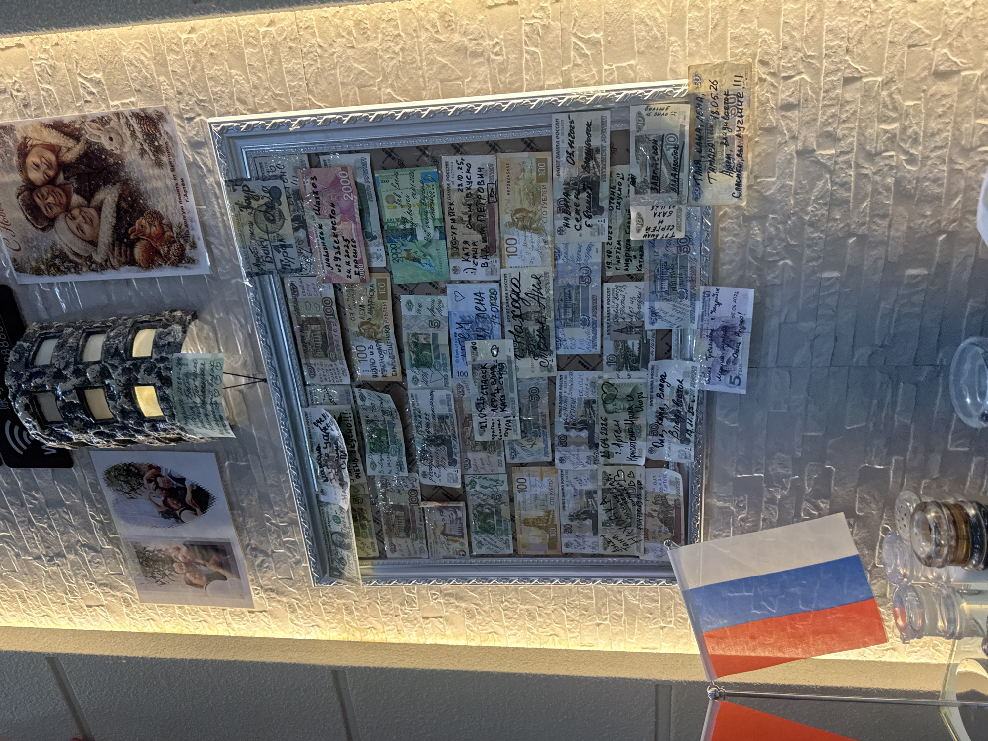 The image depicts a wall decorated with photographs and framed currency, seemingly a collection of mementos. A large framed display contains numerous banknotes, many of which have handwritten messages and dates. Above this frame, two smaller photographs are affixed to the wall, showing groups of people. To the right of these, a larger holiday-themed postcard features three individuals and animals in a snowy setting. The wall itself has a textured, light-colored finish. In the foreground, partially visible at the bottom of the image, are two flags, one appearing to be the Russian flag, and several glass containers, possibly salt and pepper shakers.
The presence of the Russian flag and the Cyrillic script on the banknotes and photographs strongly suggests a connection to Russia. Suifenhe, China, is a border city with Russia, known for its trade and tourist activity involving Russian citizens. This context suggests the location might be a business or establishment frequented by Russians, where they leave these monetary tokens as souvenirs or expressions of gratitude. The style of the decorations and the personal inscriptions point towards a casual, communal space where patrons might leave their mark.