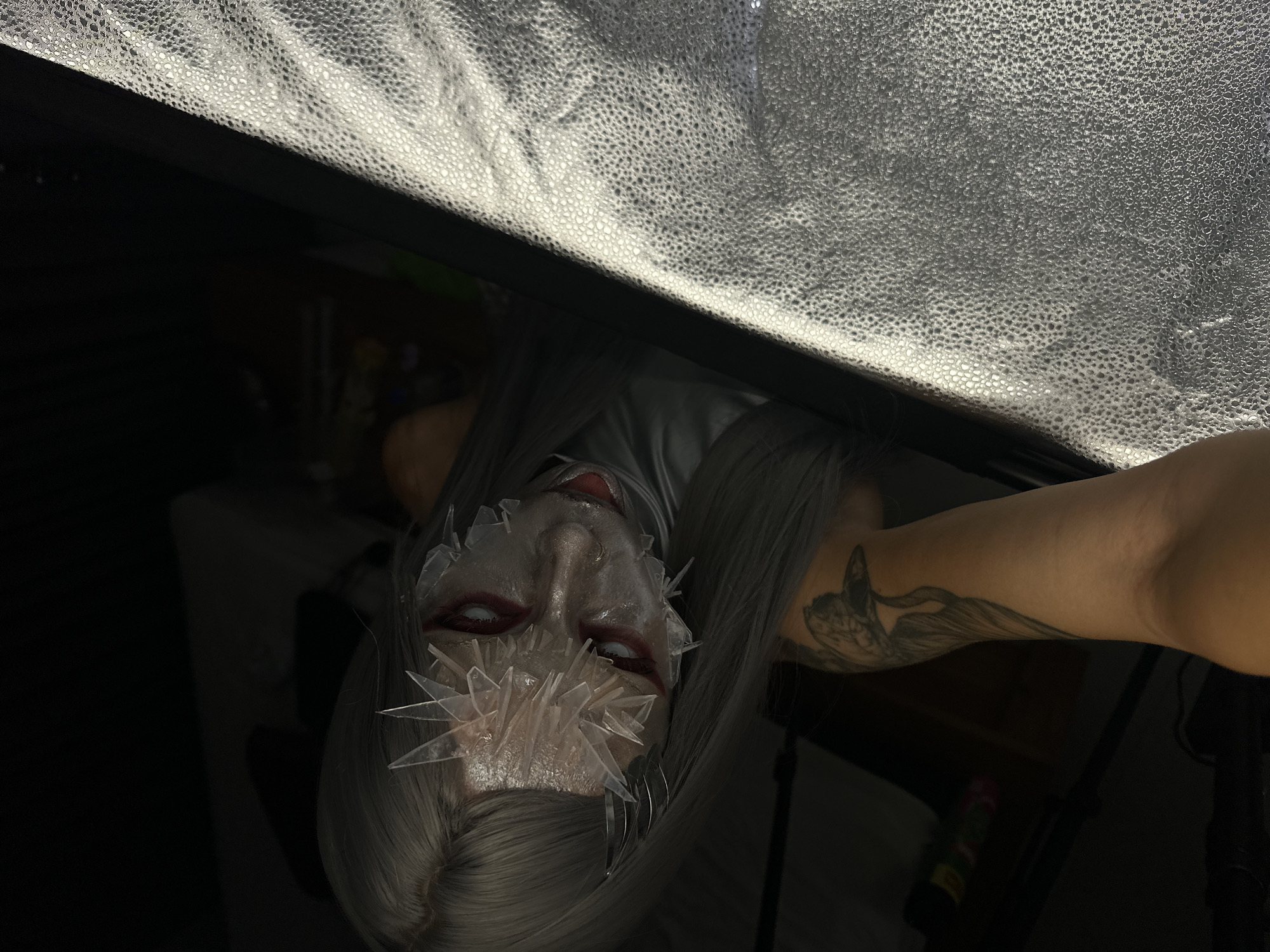 An overhead shot captures a person with striking makeup and a unique costume. Their face is partially obscured by what appears to be shards of clear material, resembling ice or glass, adhered to their skin. Their eyes are a vibrant red, and their lips are also painted red. They have long, straight, silver hair that drapes around their face and shoulders. A tattoo of a bird, possibly a crane, is visible on their arm. The lighting is dramatic, with the left side of the image cast in deep shadow, while the right side is illuminated by a textured, reflective surface, possibly a photography diffuser or screen. The background is mostly dark and out of focus, hinting at an indoor setting. The image does not provide any information about the city of North Kuta, Indonesia.