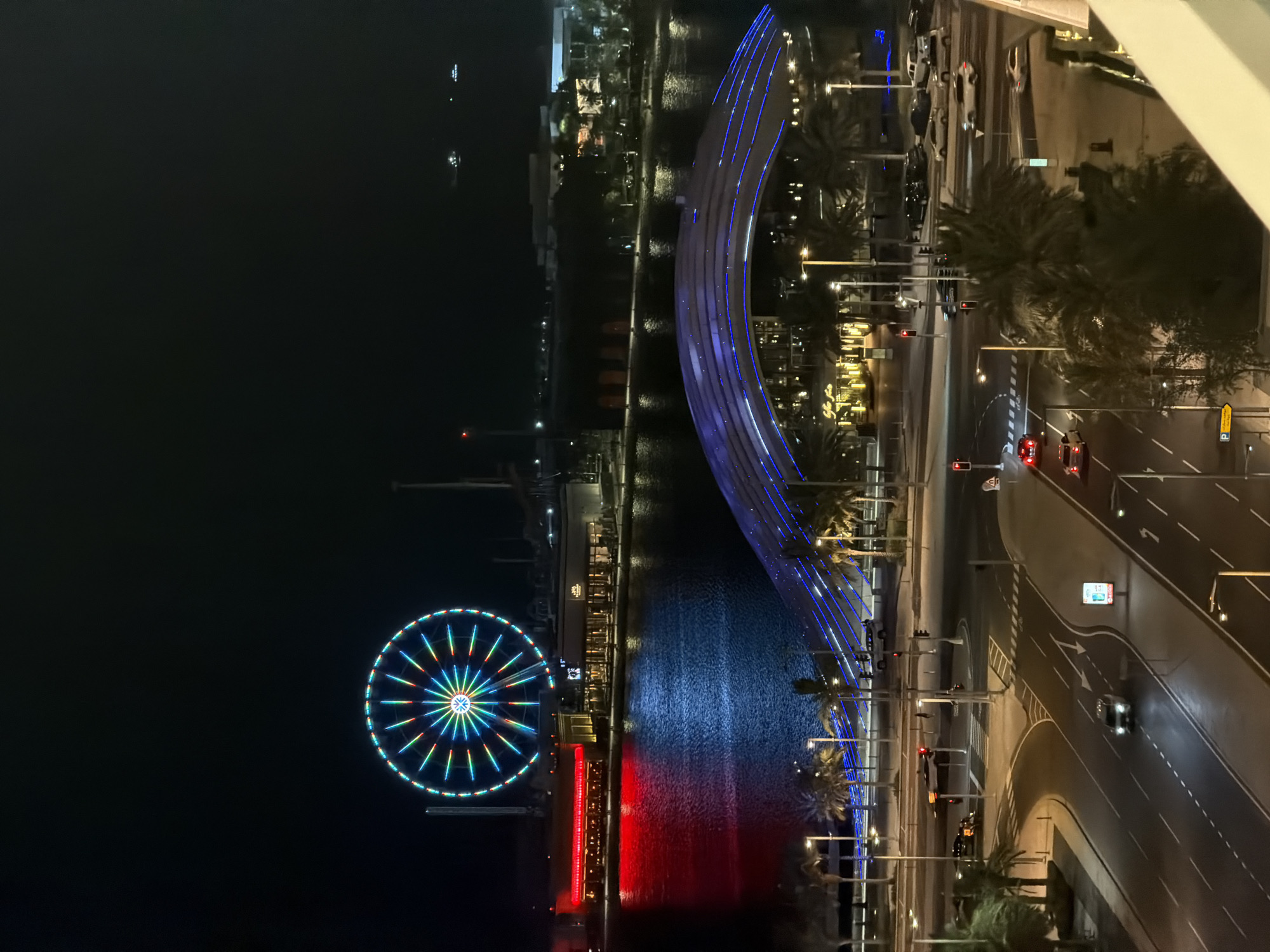 This nighttime aerial view captures a vibrant urban waterfront scene in Lusail City, Qatar. The perspective is elevated, likely from a building or balcony, overlooking a network of roads, a body of water, and prominent illuminated structures.
In the foreground, a multi-lane road intersection is visible, marked by white lines, zebra crossings, and operational traffic lights, some displaying red signals and others green. Several vehicles are present on the roads, including cars with illuminated headlights and taillights, navigating the intersection. The roads are flanked by numerous mature palm trees, illuminated by streetlights that cast bright yellow-orange light. A white railing, indicative of a balcony edge, is partially visible in the bottom right.
The midground features a calm body of water, likely a canal or bay, reflecting the artificial lights from the surrounding environment. Spanning this water is a distinctive pedestrian bridge, characterized by multiple parallel arches with integrated blue strip lighting that creates a luminous, segmented effect. Along the far side of the water, several commercial buildings or restaurants are brightly lit, with their warm interior and exterior lights reflecting predominantly red and blue hues onto the water's surface. Palm trees also line this waterfront.
Dominating the background on the left is a very large Ferris wheel, brilliantly illuminated with a complex pattern of multi-colored lights including blue, green, red, and yellow, radiating from its central hub to the outer rim. To the right of the Ferris wheel and across the water, other distant urban structures and faint lights are discernible against the dark, clear night sky.